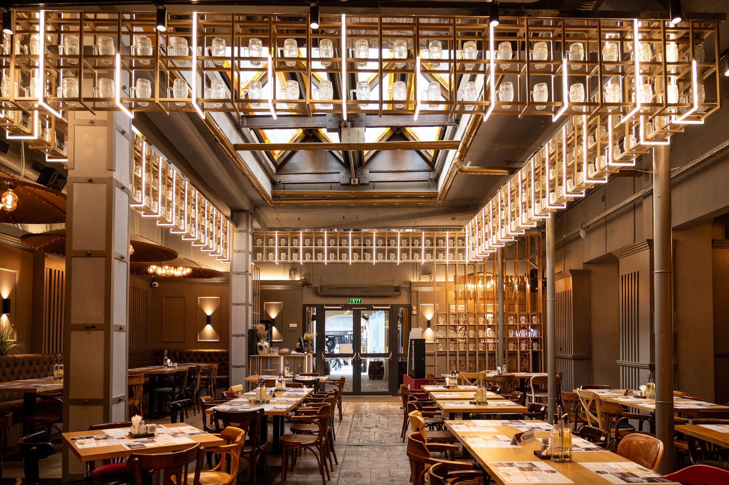 Interior of a restaurant with wooden tables and chairs, modern lighting fixtures, decorative shelves with bottles, and a glass double door at the entrance.