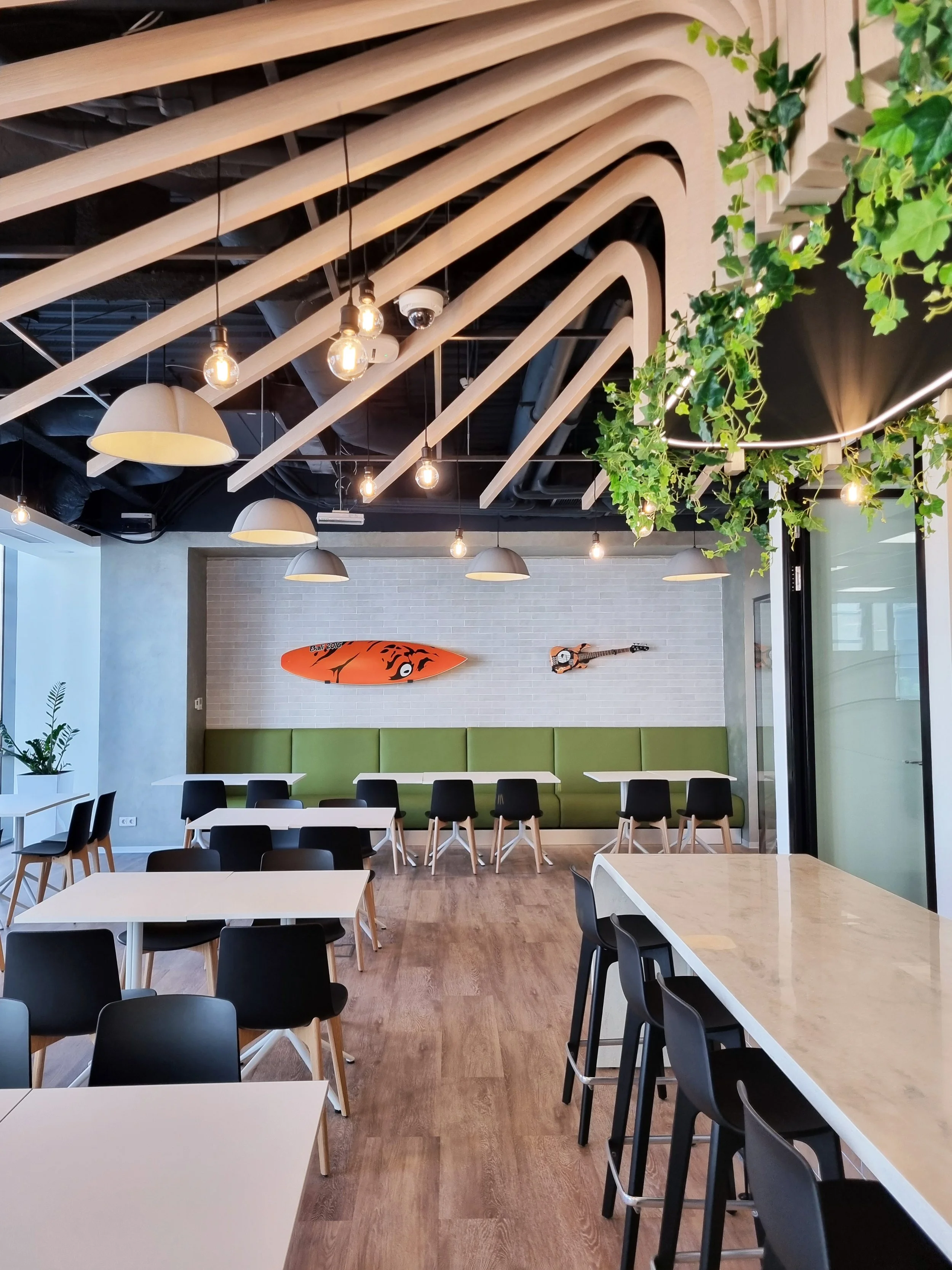 Modern restaurant interior with white tables, black chairs, green upholstered bench, decorative surfboard and guitar wall art, hanging light bulbs, wooden ceiling beams, and green plant hanging from ceiling.