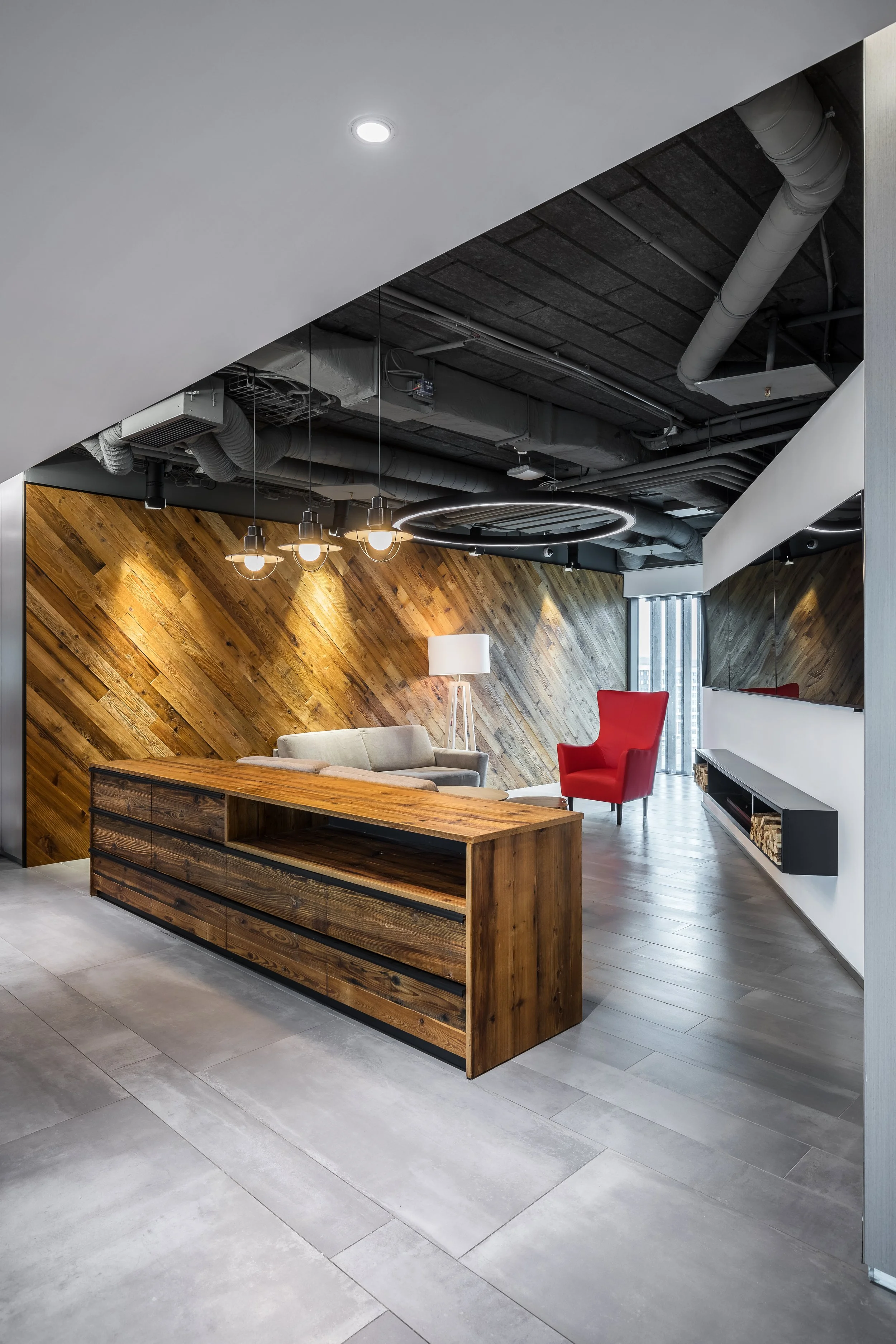 Modern office lounge with a wooden wall, a beige sofa, a red armchair, a floor lamp, and hanging light fixtures.