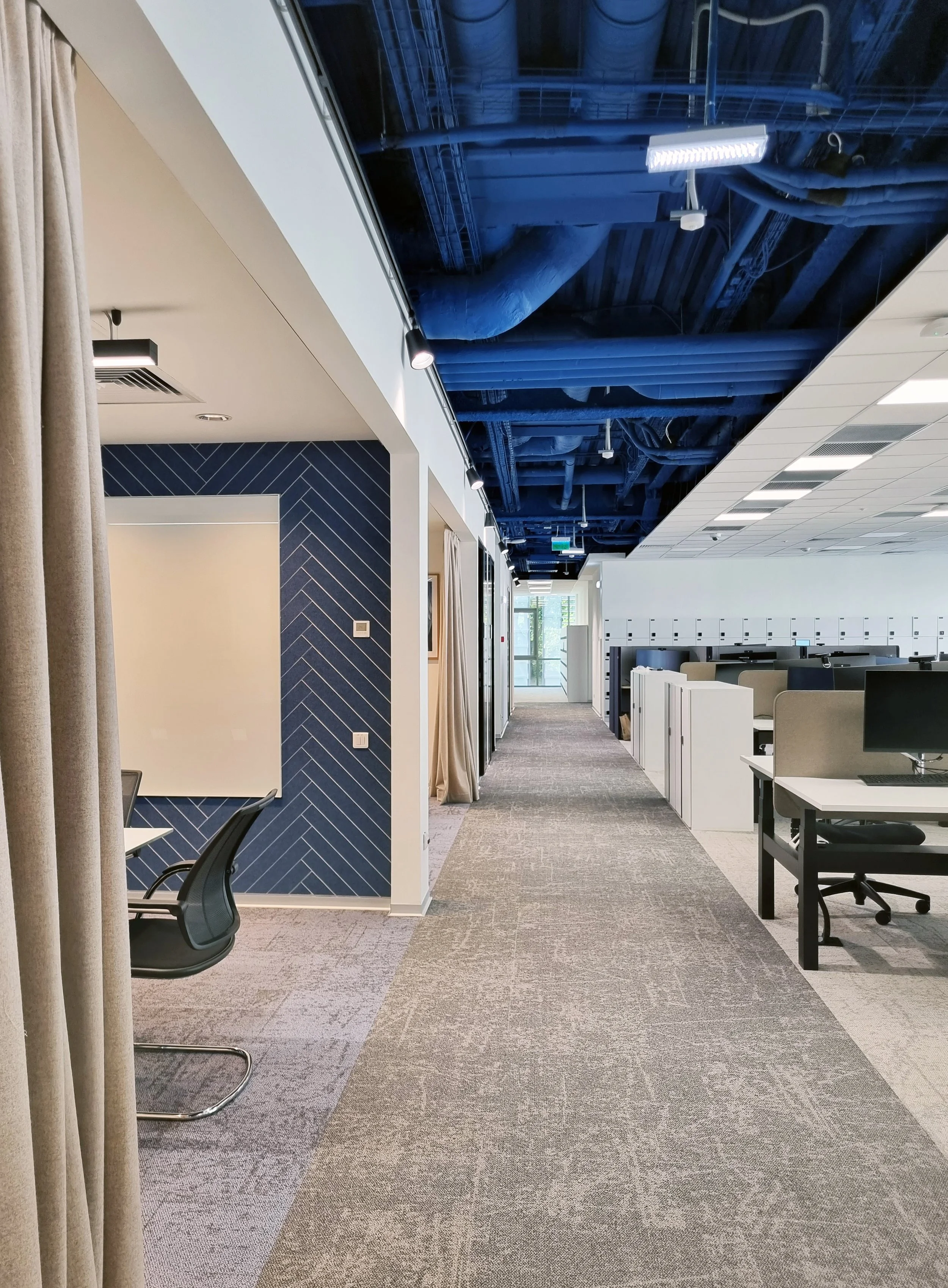 Modern office space with workstations, cubicles, a blue accent wall, and an exposed blue ceiling with ductwork and lighting fixtures.