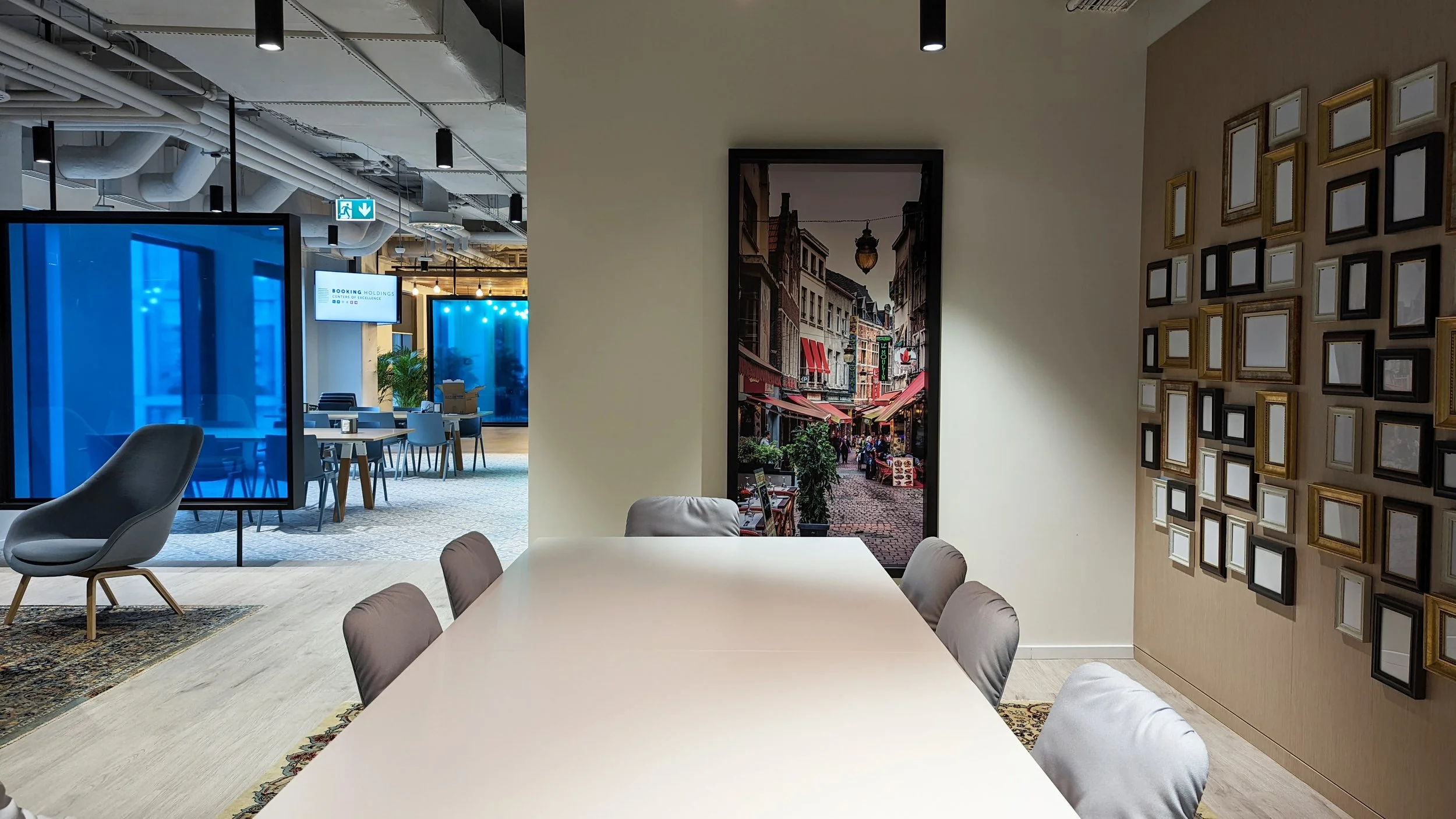 An interior conference room with a long white table, surrounded by gray upholstered chairs, and decorated with a gallery wall of empty picture frames. A large vertical photograph of a European street scene hangs on the wall. In the background, a modern open office space with blue accents, tables, and seating can be seen.