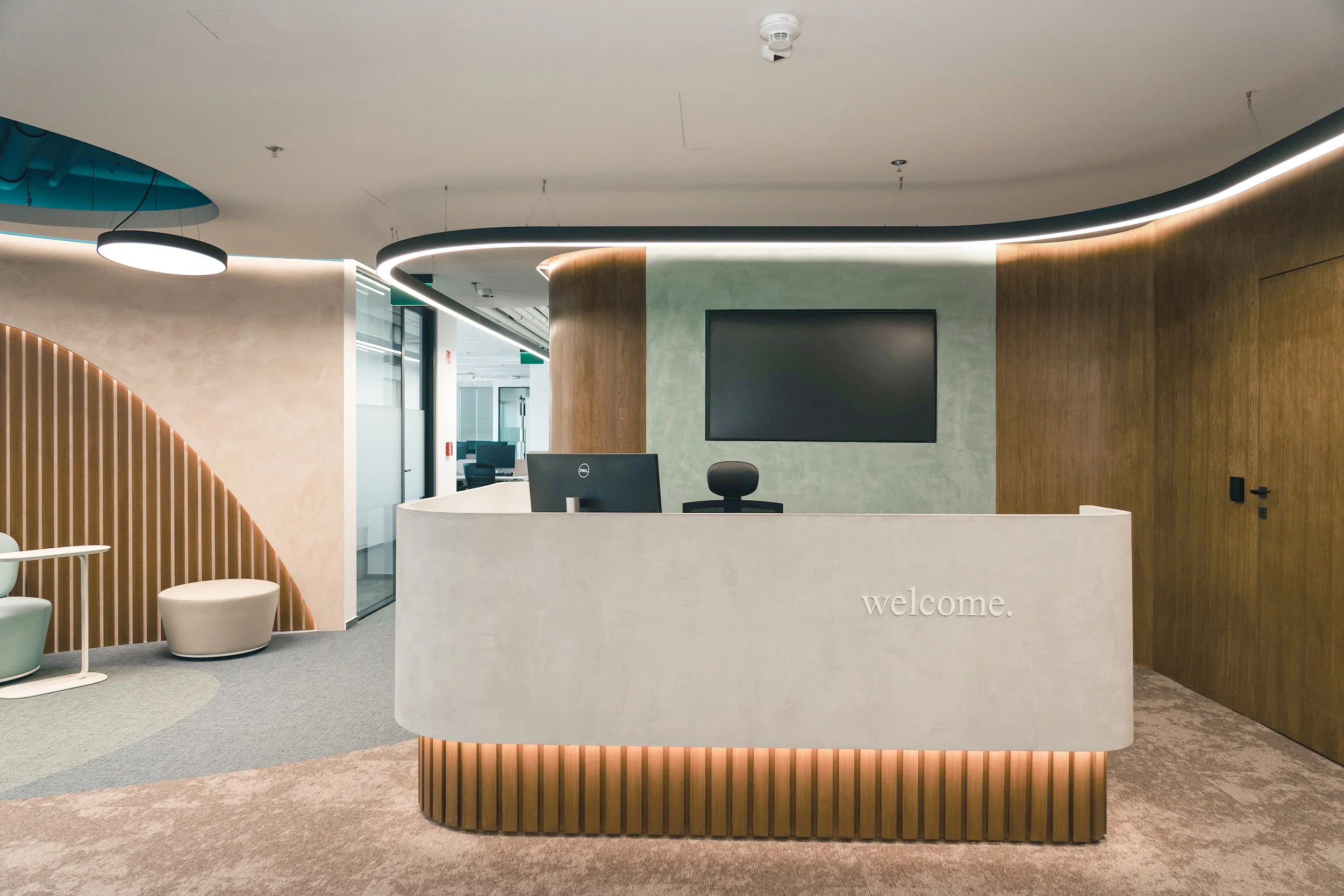 Modern office reception area with white curved desk, computer, wall-mounted TV, and seating area with chairs and ottoman