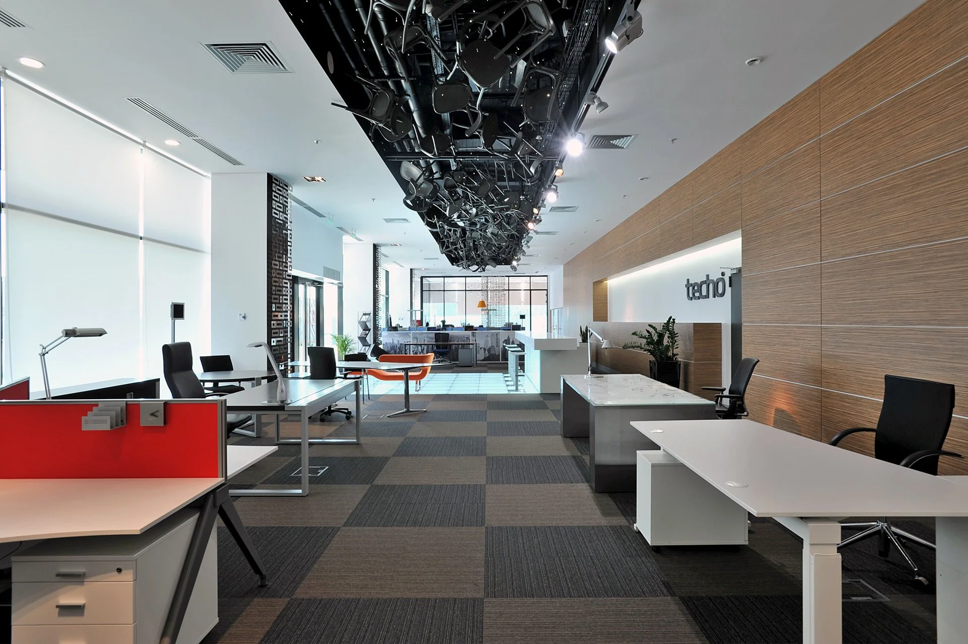 Modern office space with desks, chairs, and a reception area, illuminated by natural light through large windows, and decorated with plants and wood paneling.