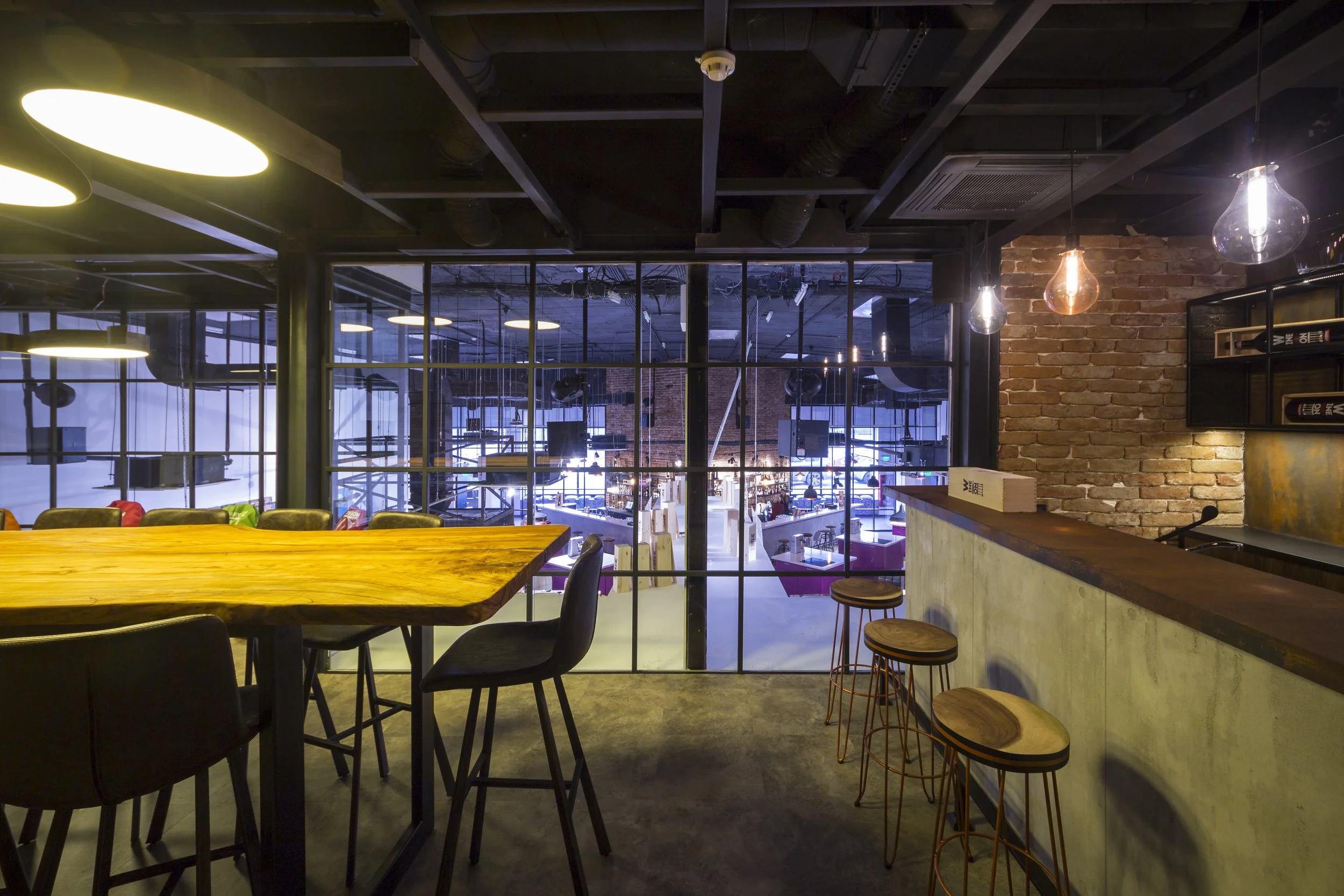 Interior of a modern restaurant or cafe with a high wooden table, black chairs, and bar stools. Exposed brick wall with hanging glass light fixtures. Large glass window revealing a seating area with colorful chairs and tables.