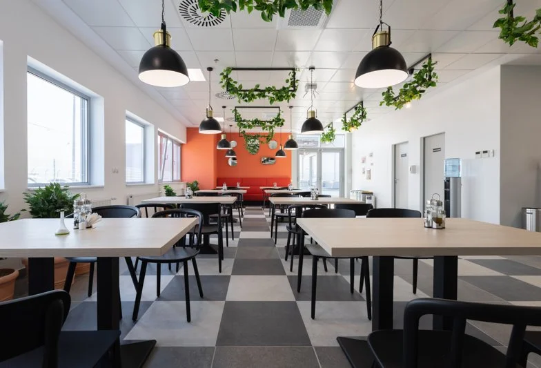 Empty restaurant with white walls, black pendant lights, green hanging plants, and black chairs around light wood tables. Large windows on the left and red accent wall at the back.