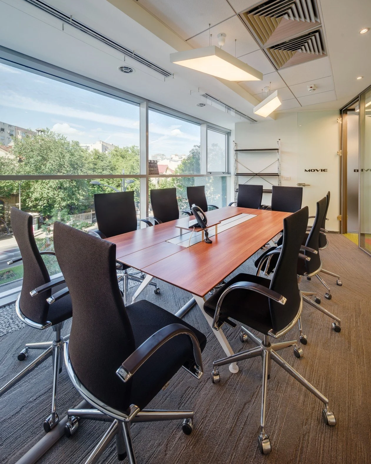 A modern conference room with a large wooden table surrounded by nine black office chairs, large windows with a city view, and ceiling lights.