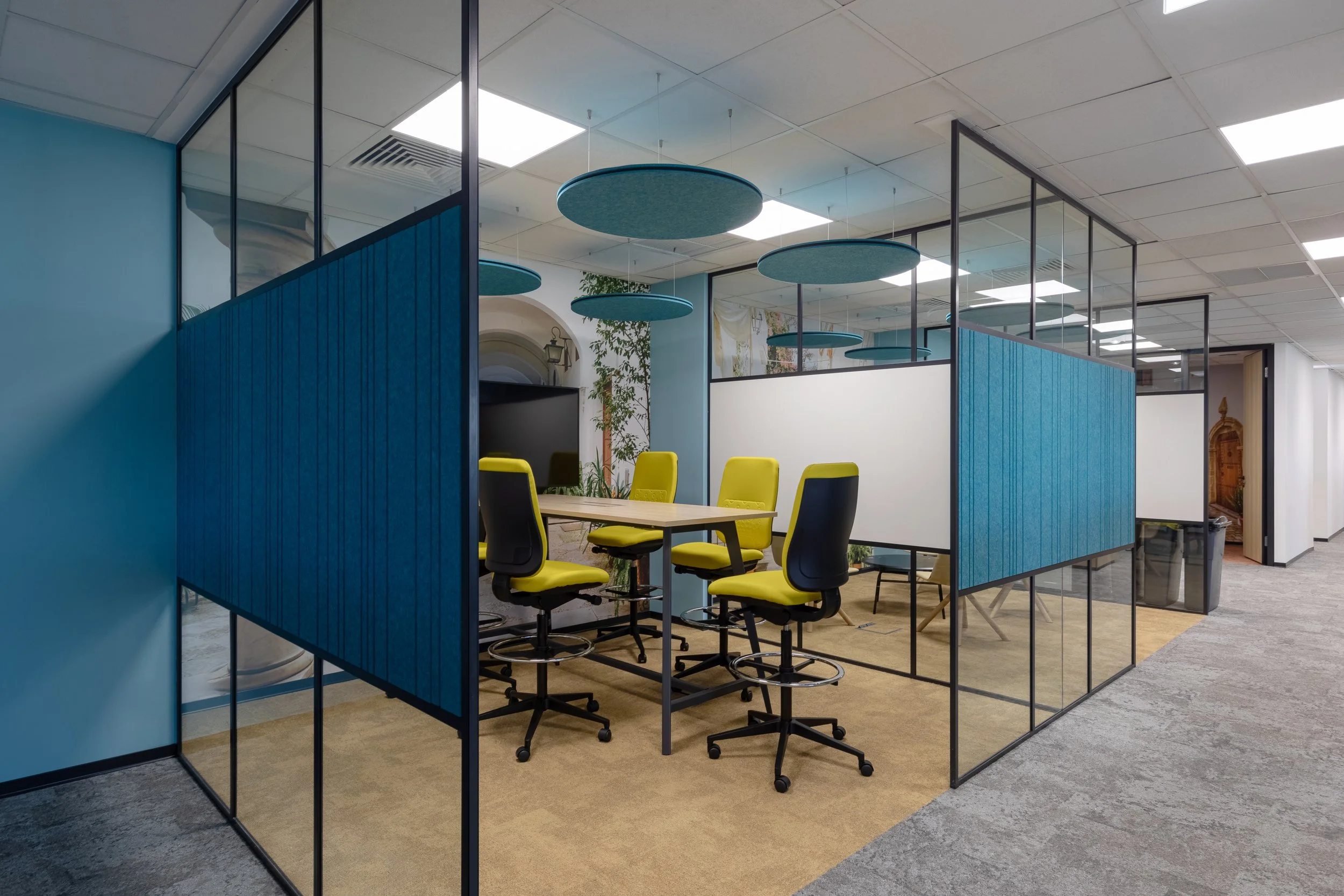 Modern office meeting room with a rectangular table surrounded by six yellow and black chairs, enclosed by glass walls with blue and white accents, ceiling lights, and hanging blue acoustic panels.