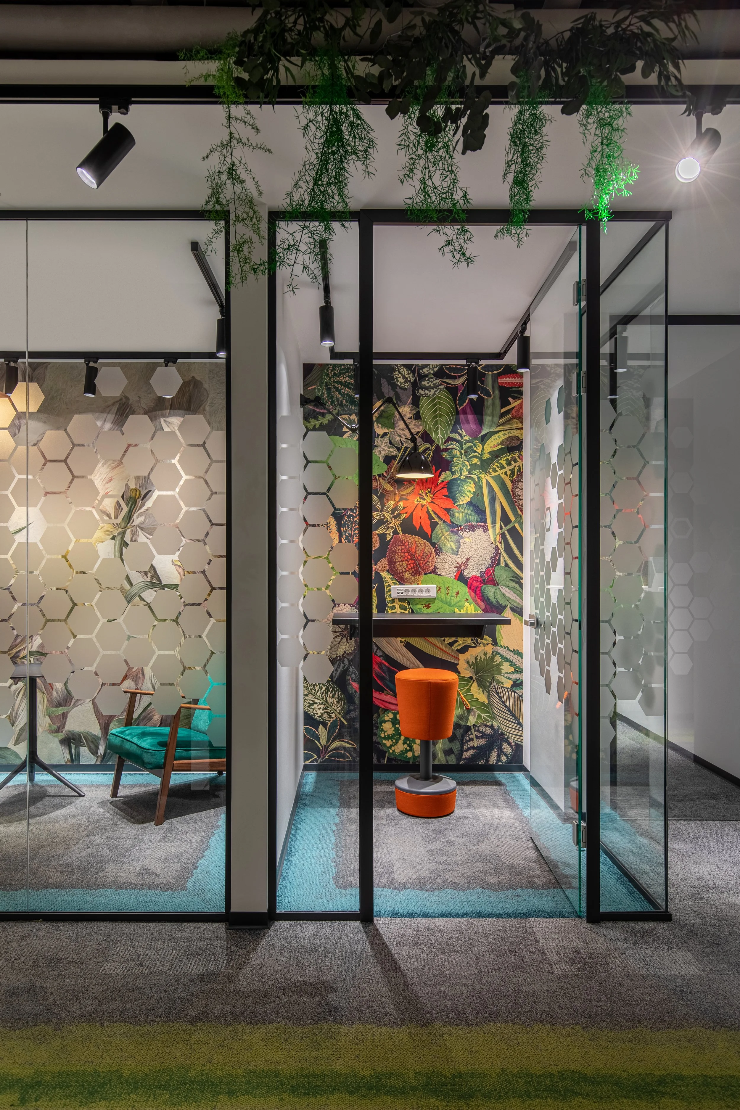 Modern office with glass partition walls, colorful botanical wallpaper, and contemporary furniture including a green lounge chair and orange stool.