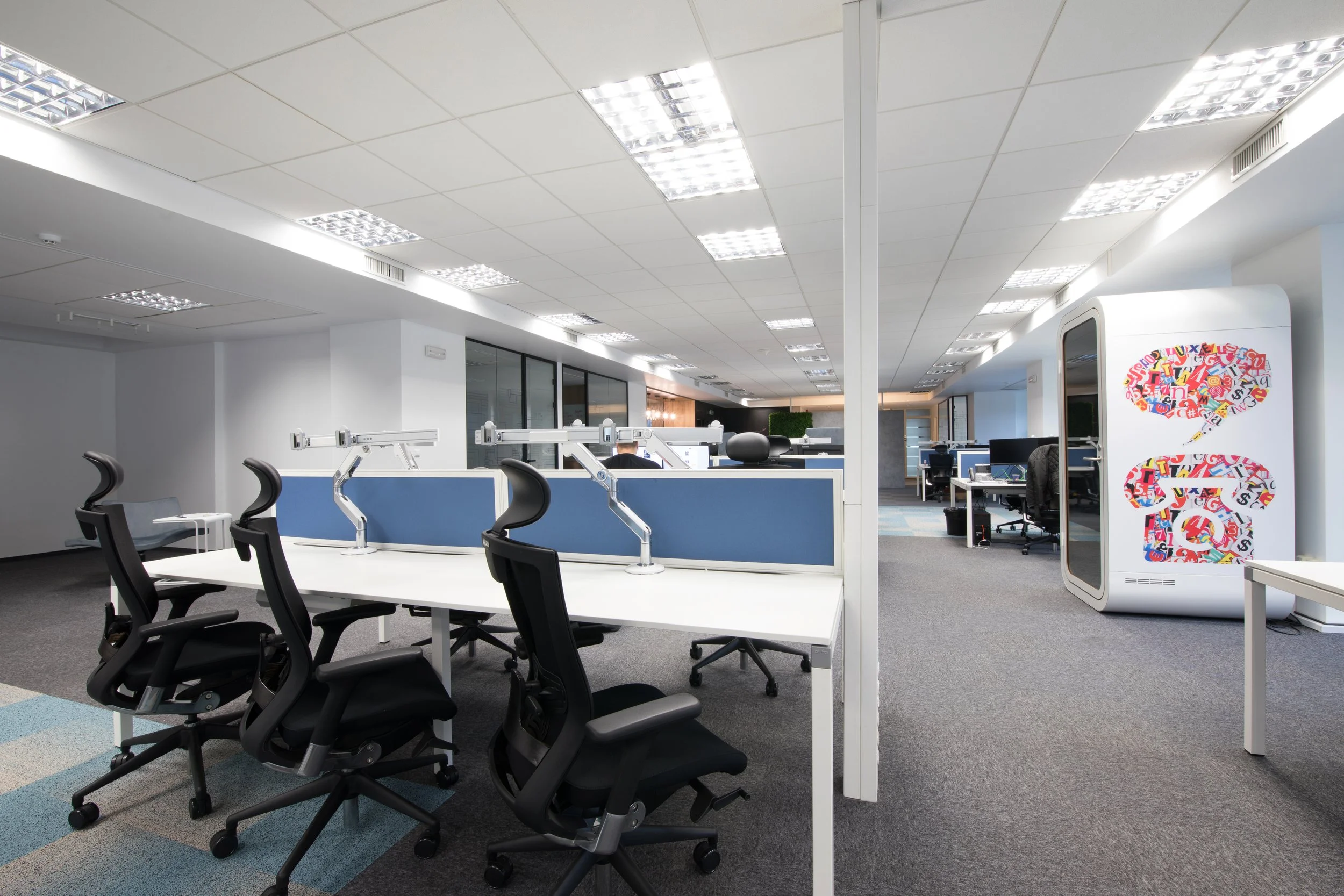 Empty modern office workspace with black ergonomic chairs around white desks, blue partitions, computers, and a colorful recycling/garbage bin with a speech bubble design, in a well-lit environment.