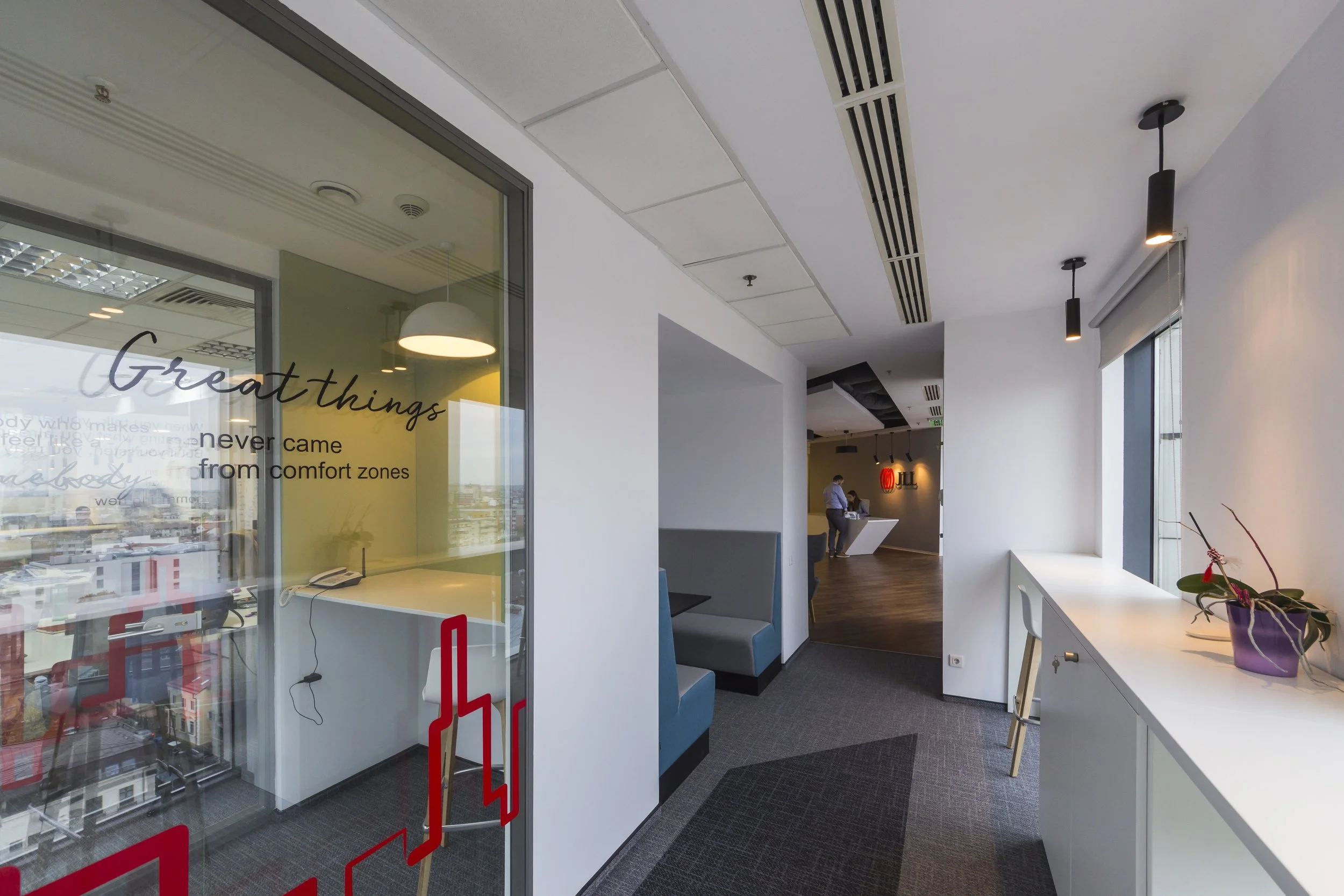 Modern office space with a glass door, white walls, gray and blue seating booths, a window with a flowerpot on a white counter, and a reception desk in the background with two people standing nearby.