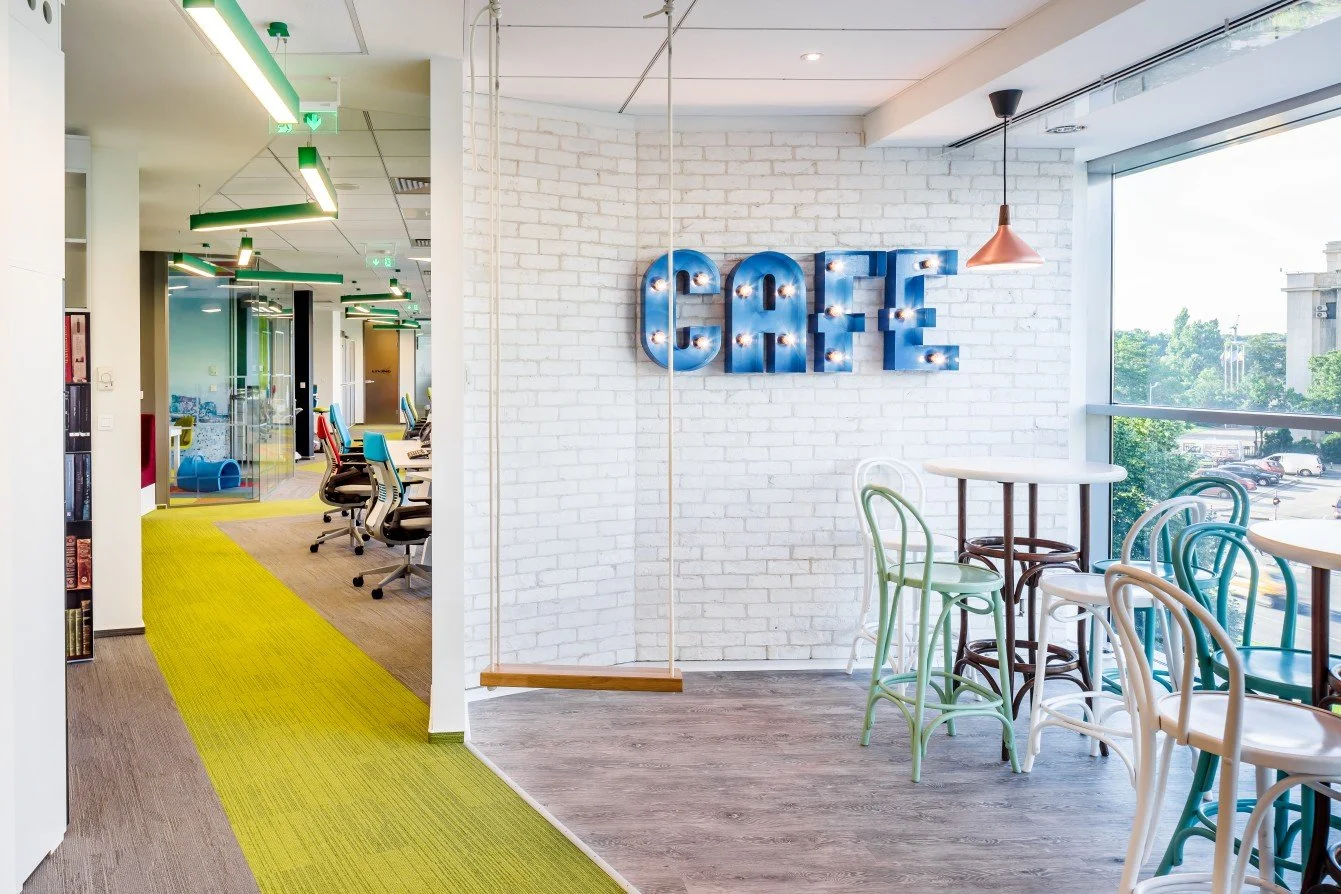 Bright modern office space with a white brick wall featuring a lighted blue "CAFE" sign, tables and chairs near a large window showing outdoor parking and trees.