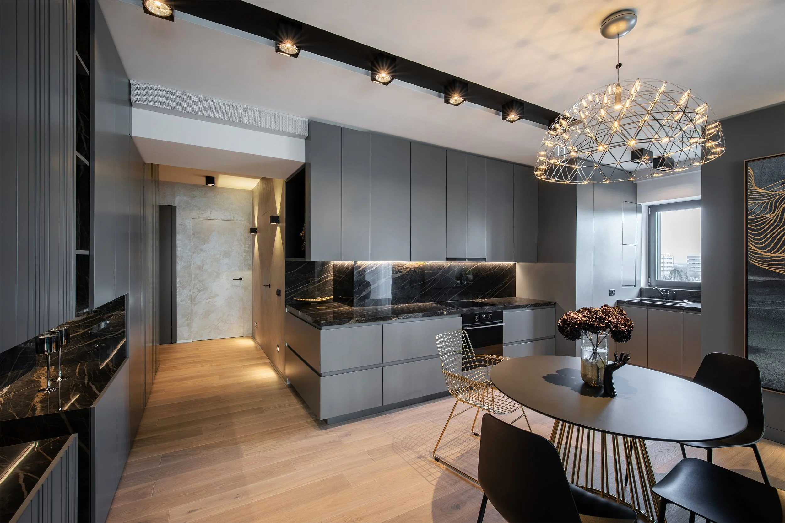Modern kitchen with gray cabinets, black marble countertops, and a round dining table with black and gold chairs, illuminated by an artistic chandelier and recessed ceiling lights.