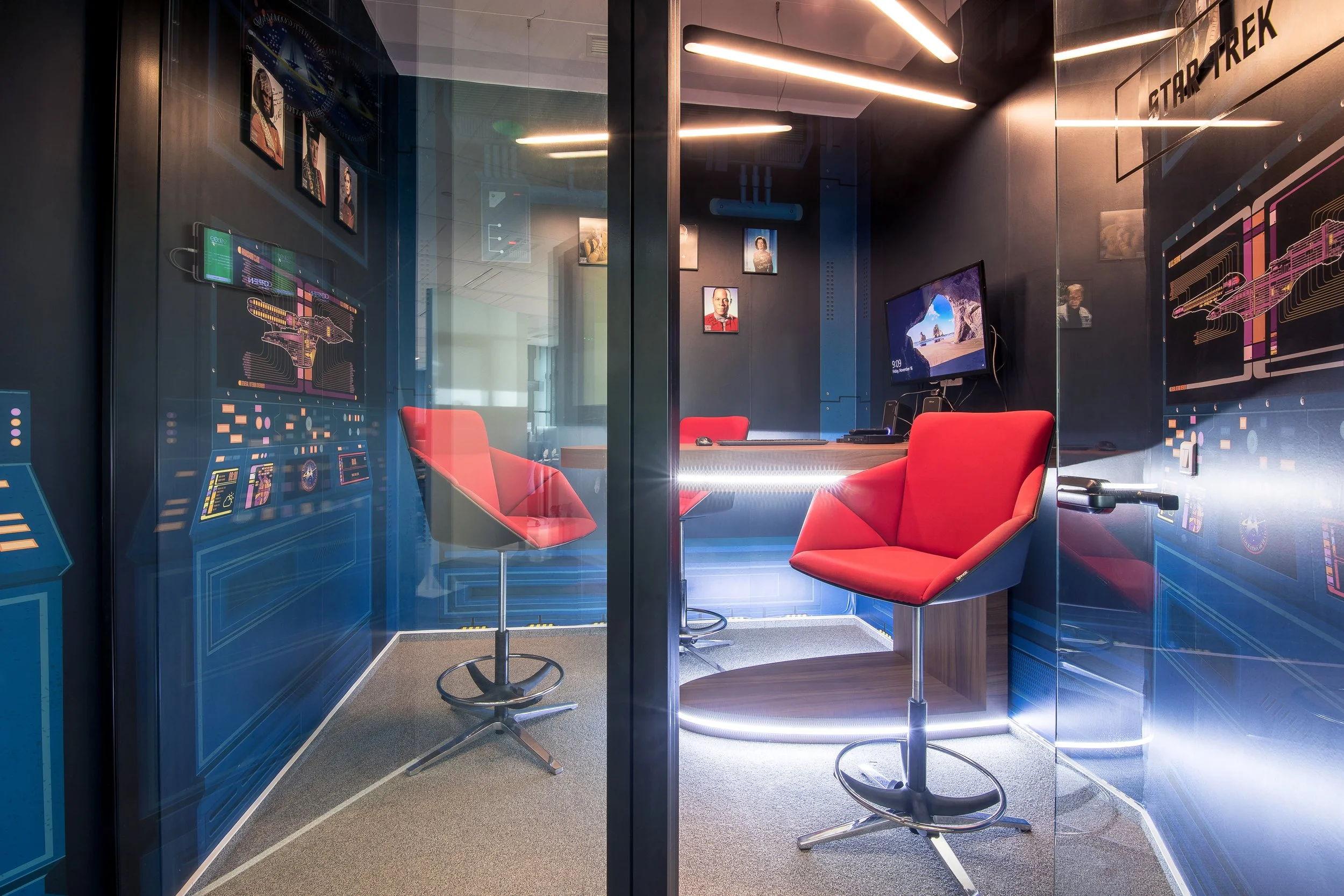 A modern office or conference room with dark walls decorated with Star Trek-themed artwork and framed photos. The room has a glass wall or door, red chairs with metallic bases, a desk with a computer monitor showing a desert landscape, and futuristic lighting strips on the ceiling and along the floor.