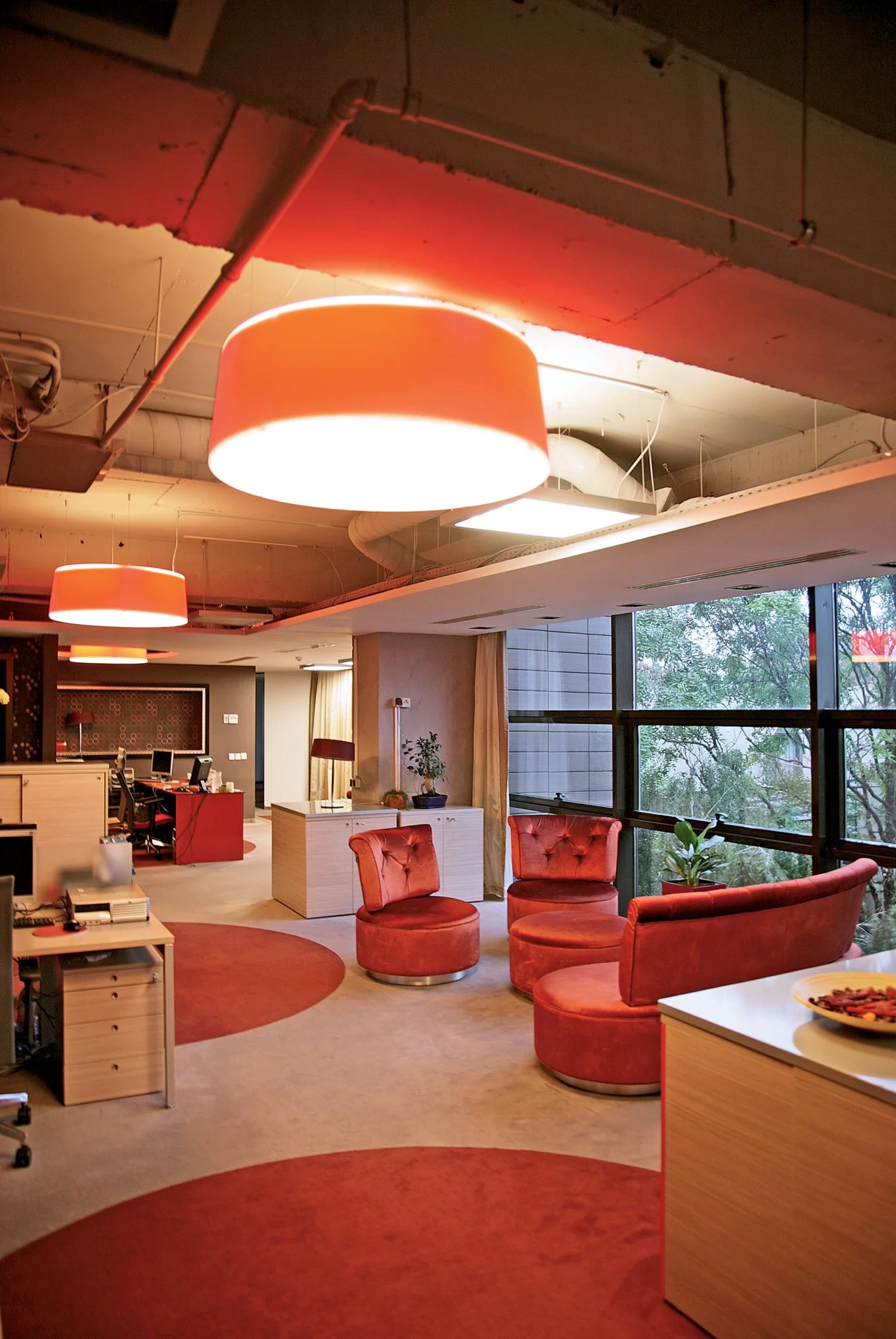 Interior office space with large orange ceiling lights, pink velvet chairs, and large windows showing trees outside.