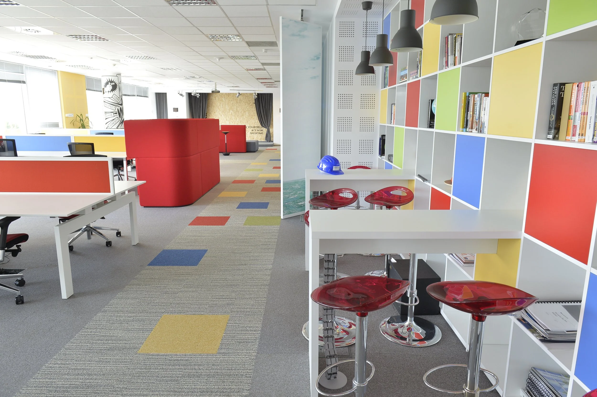 Modern, colorful indoor office space with open layout, white desks with black chairs, red semi-private sitting areas, patterned carpet with yellow, blue, red, and gray tiles, a bookshelf with books and a blue safety helmet, and hanging black pendant lights.