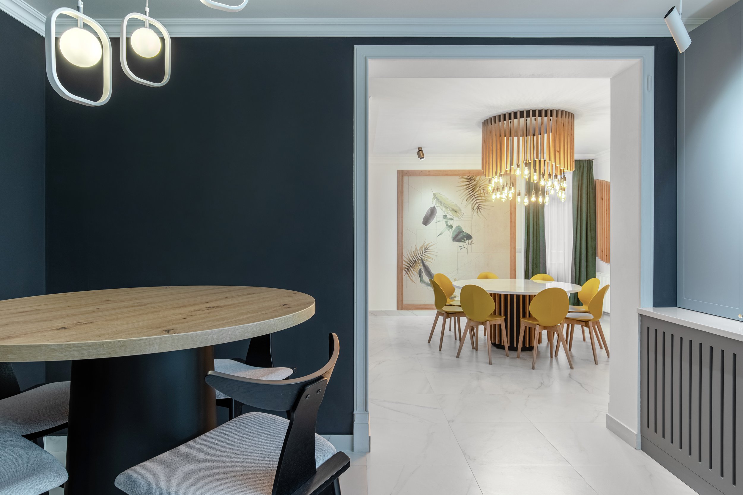 Modern dining room with a round white table and yellow chairs, separated by a doorway from a darker room with a small round wooden table and two chairs, and a contemporary chandelier with wooden slats and hanging light bulbs.