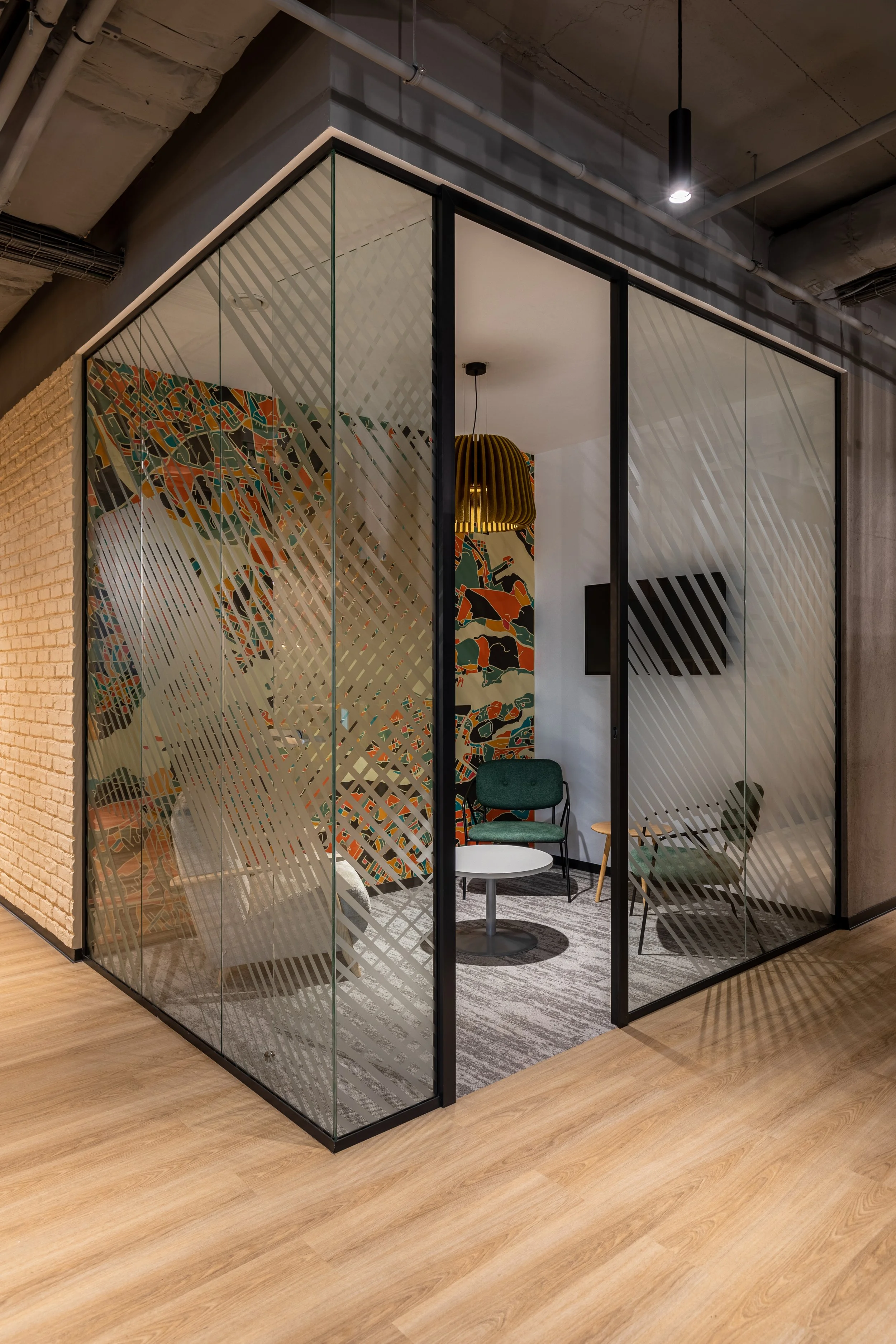 Modern glass-walled conference room with colorful abstract wallpaper, green chairs, a small round table, and a black flat-screen TV on the wall. Exterior has wood flooring and exposed ceiling pipes.
