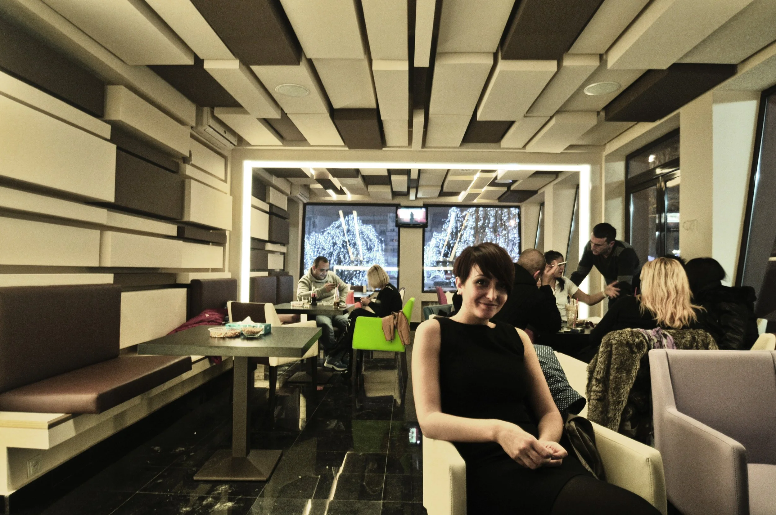 People dining in a modern restaurant with a woman smiling at the camera in the foreground, others seated at tables behind, large windows with snow outside, stylish interior design.