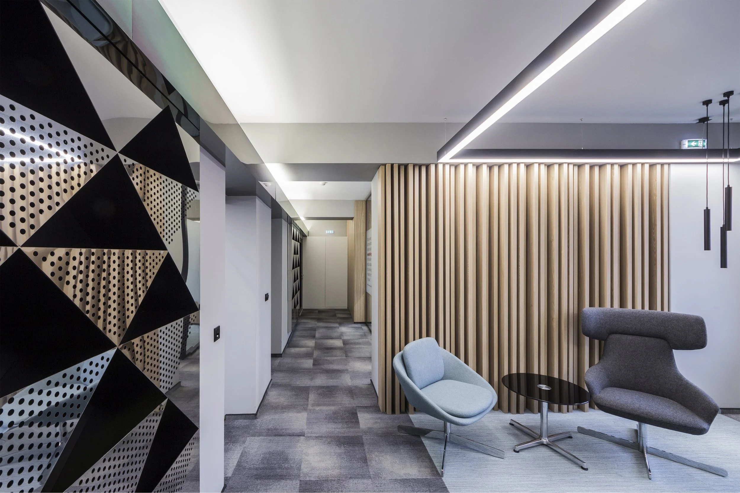 Modern interior of an office lounge area with two stylish chairs, a small round table, a wooden vertical slat wall, and contemporary lighting