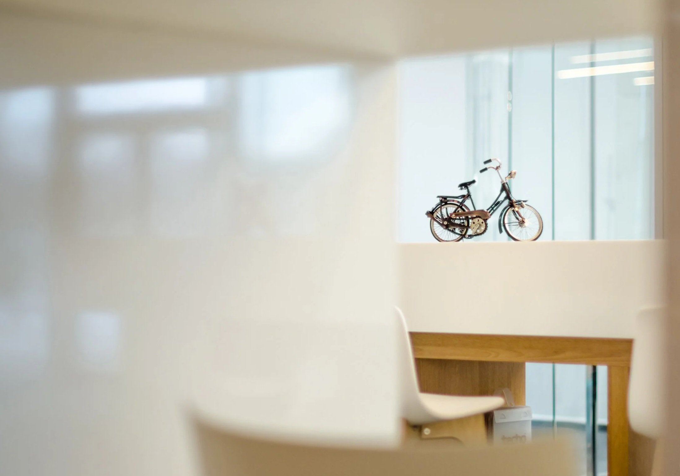 Model of a bicycle on a balcony or window ledge, viewed through an opening in the wall, with glass windows in the background in an indoor space.