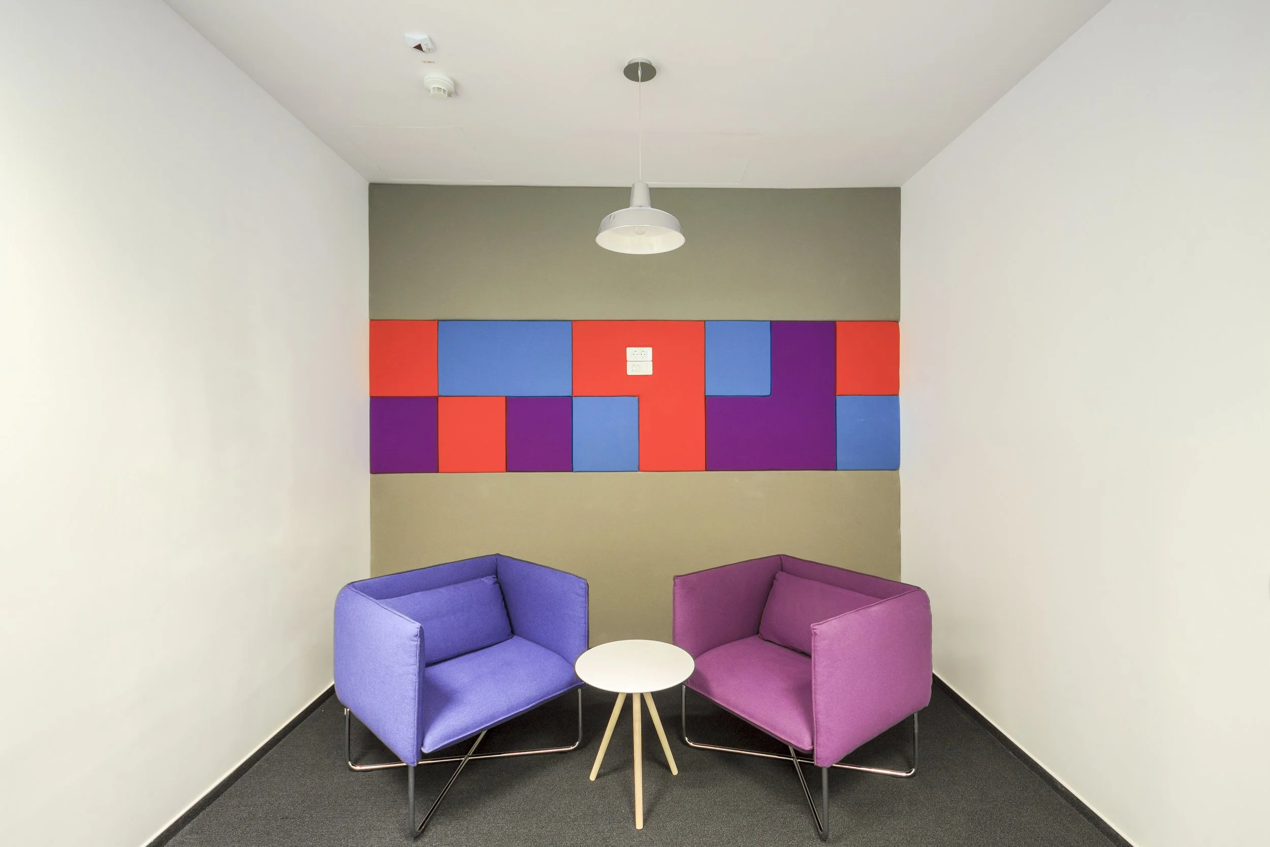 A small, modern office waiting area with two colorful armchairs, a white round table between them, a geometric patterned wall in the background, and a ceiling light.