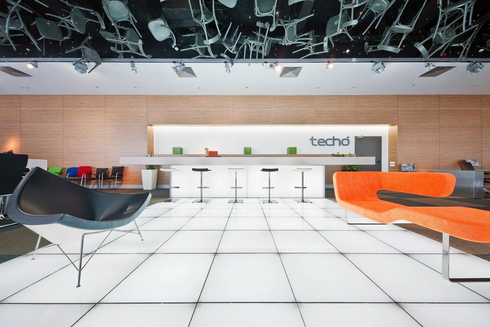 Modern office lobby with white tile floor, wooden wall panels, and a white reception desk. Black, white, and colorful chairs are arranged around the space. Ceiling has black panels with chairs hanging upside down. The word 'techo' is displayed on the wall behind the reception desk.