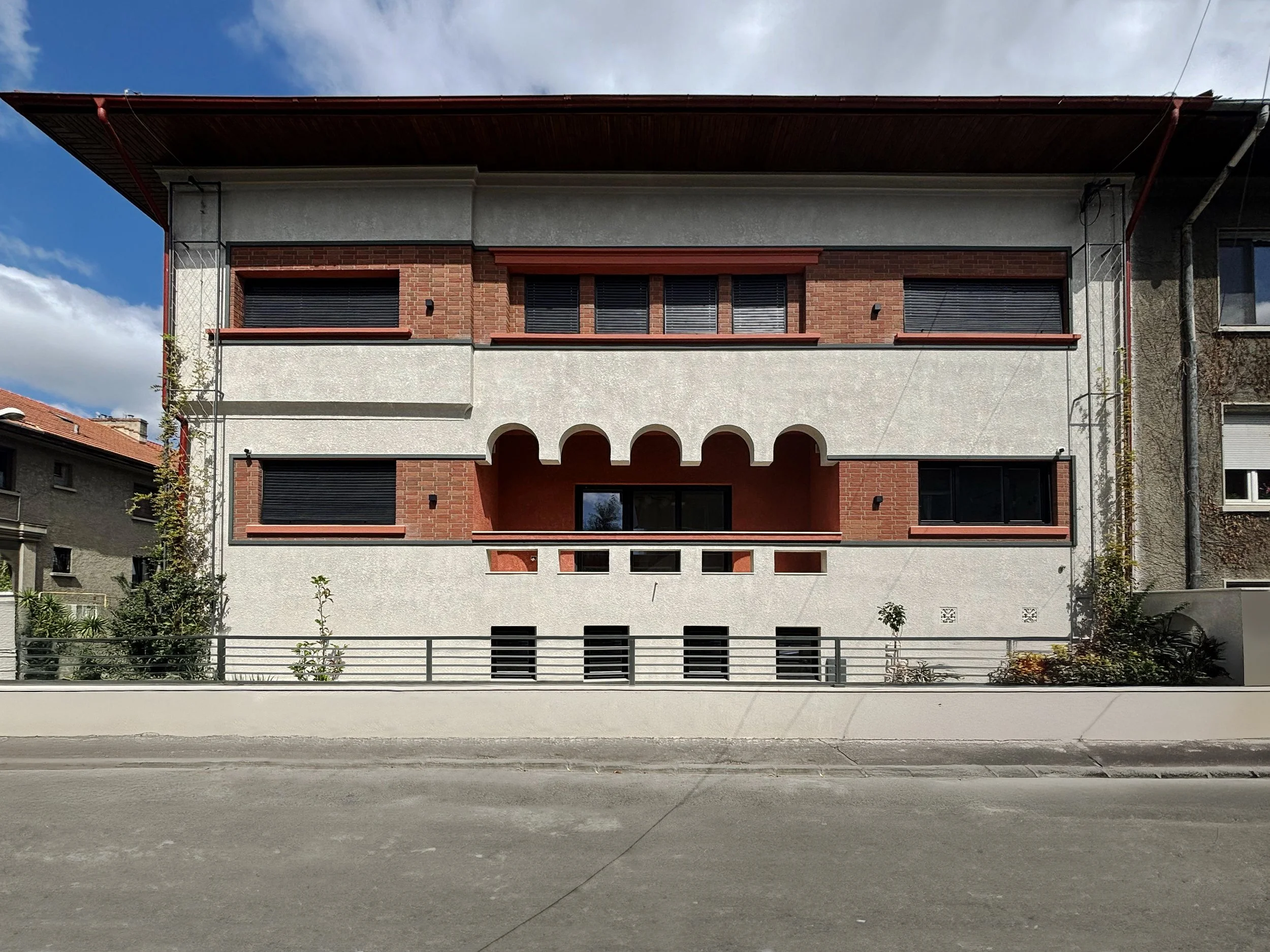 Modern, multi-story residential building with white and red brick exterior and arched balcony at center, set on a street with a concrete sidewalk and railing.