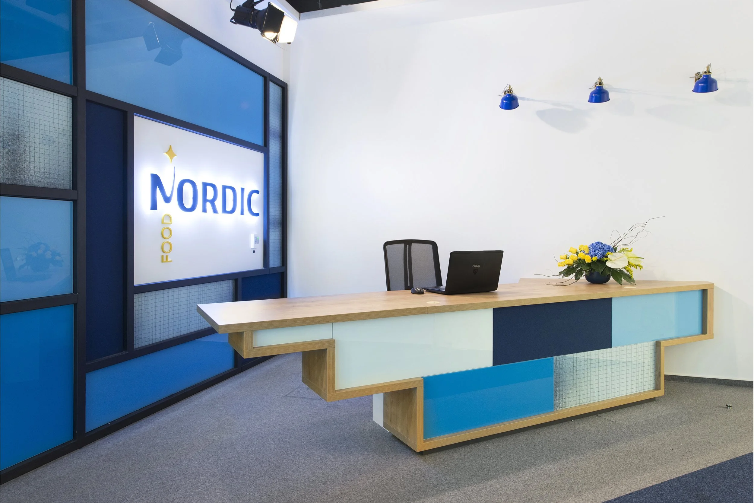 Office reception area with a wooden desk, a black office chair, a laptop, a mouse, a decorative flower arrangement with yellow and blue flowers, blue wall-mounted lights, and a large illuminated sign that reads 'Nordic Food' on the wall.