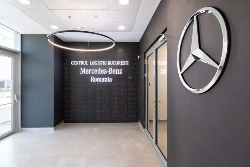 Modern office lobby with a Mercedes-Benz logo on a dark gray wall, a circular LED light fixture on the ceiling, and a sign that reads 'CENTRUL LOGISTIC BUCURESTI Mercedes-Benz Romania' near an elevator door.