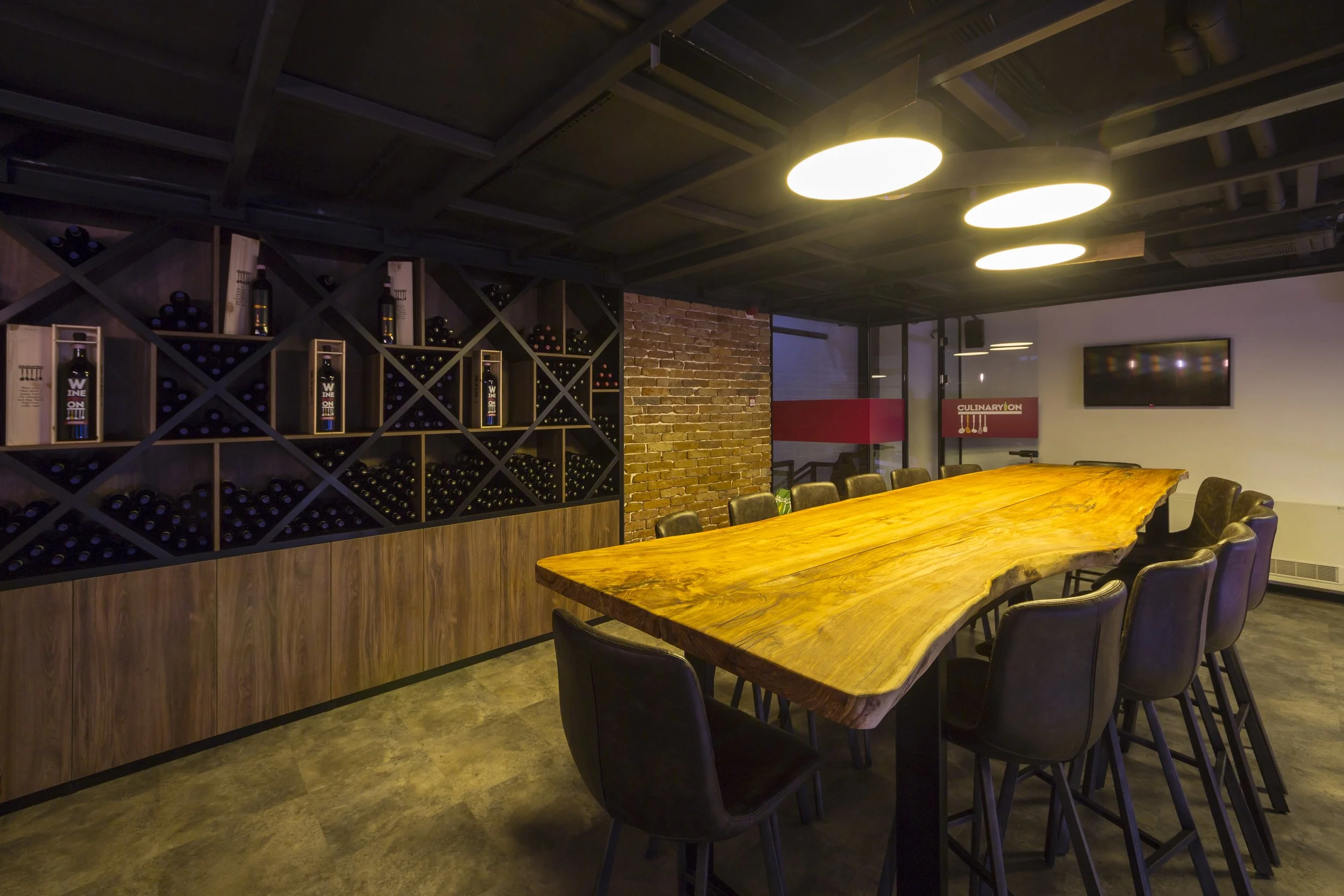 Interior of a wine tasting room with a long wooden table, black chairs, wine bottles on shelves, exposed brick wall, and a TV screen.