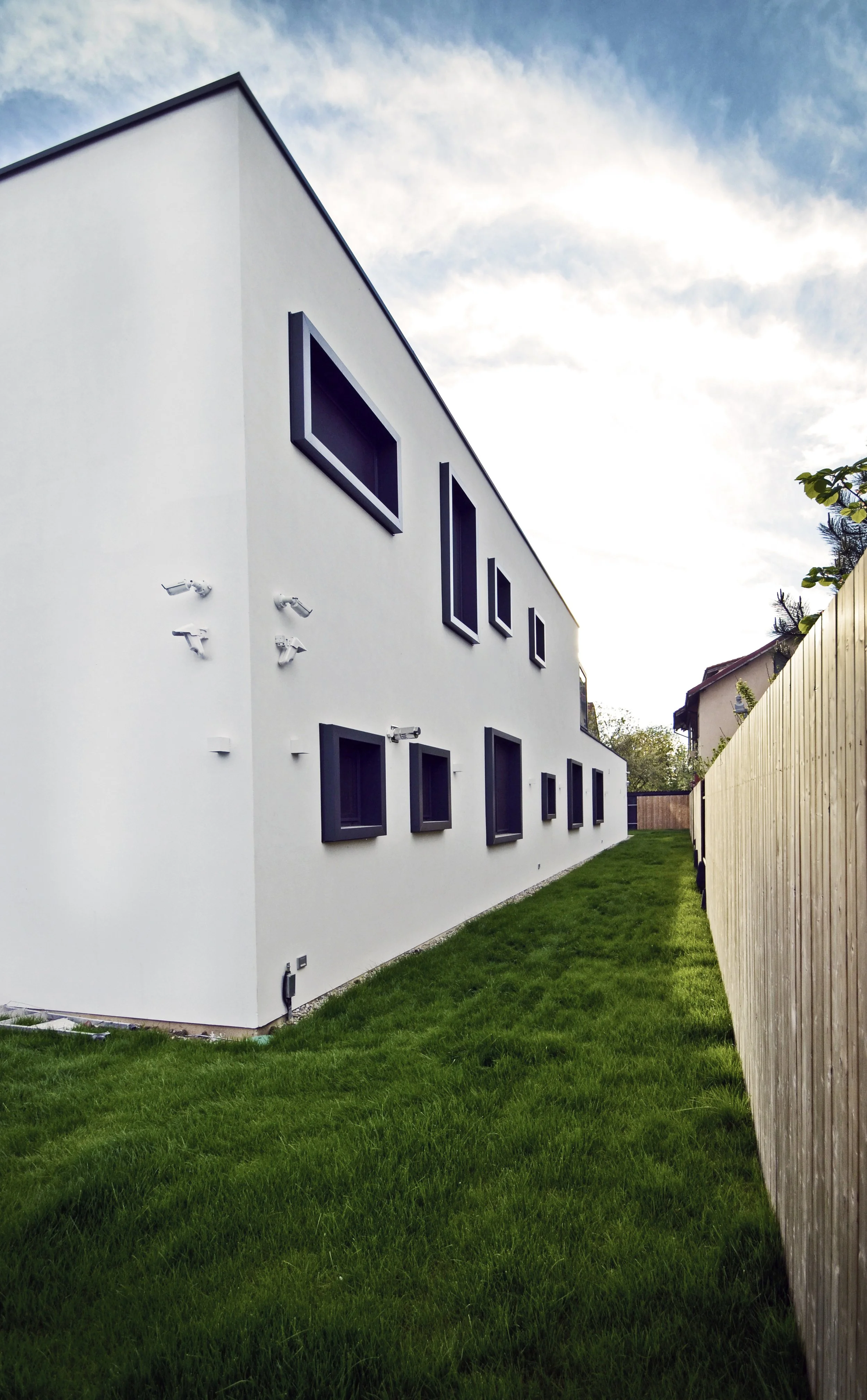 Modern white building with dark rectangular windows on a grassy lot, fenced with a wooden fence, under a cloudy sky.