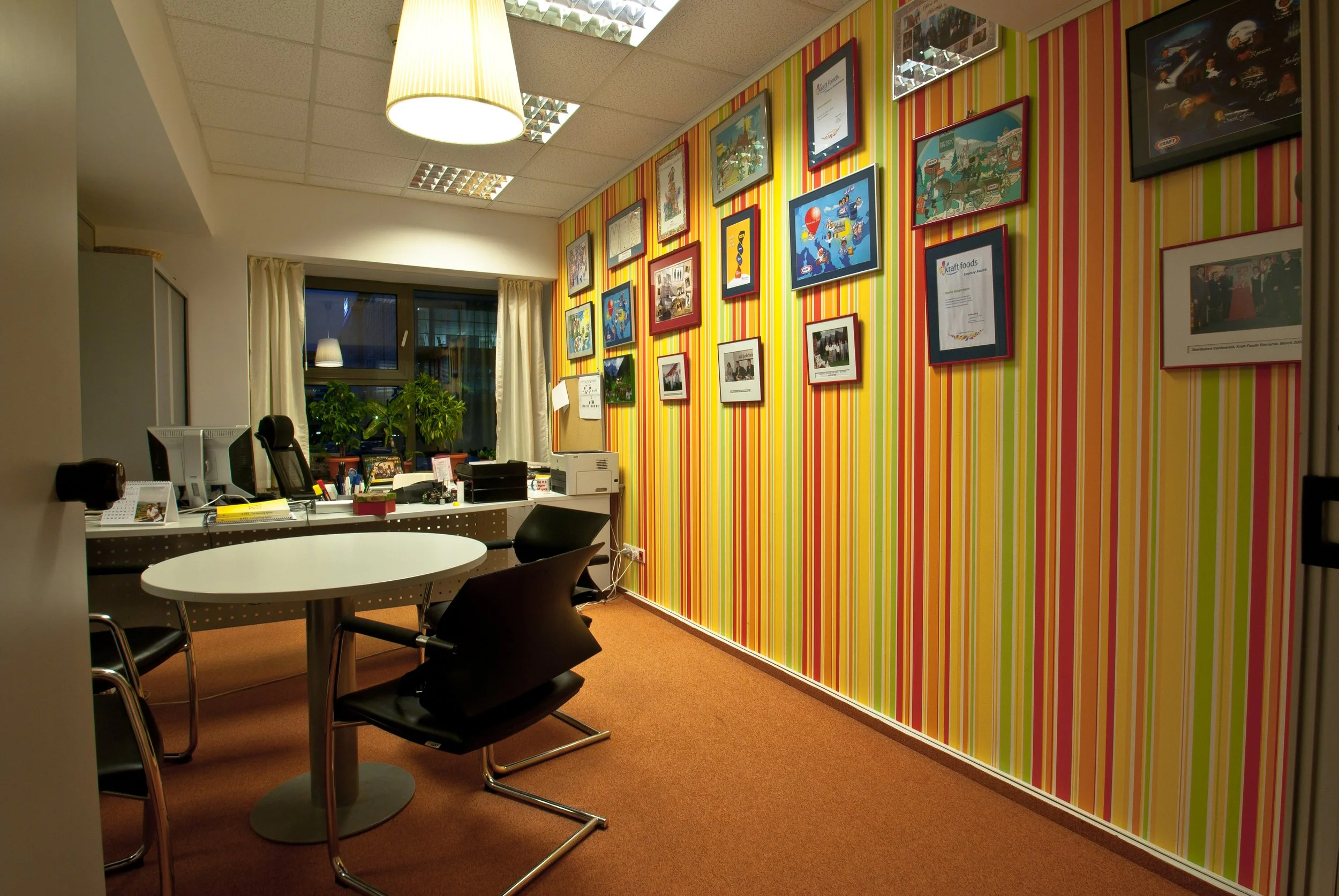 Office with a striped yellow, orange, and green wall adorned with framed pictures and awards. A desk with three chairs, a computer, plants, and office supplies is situated near a window with cream curtains. Overhead lighting illuminates the room.
