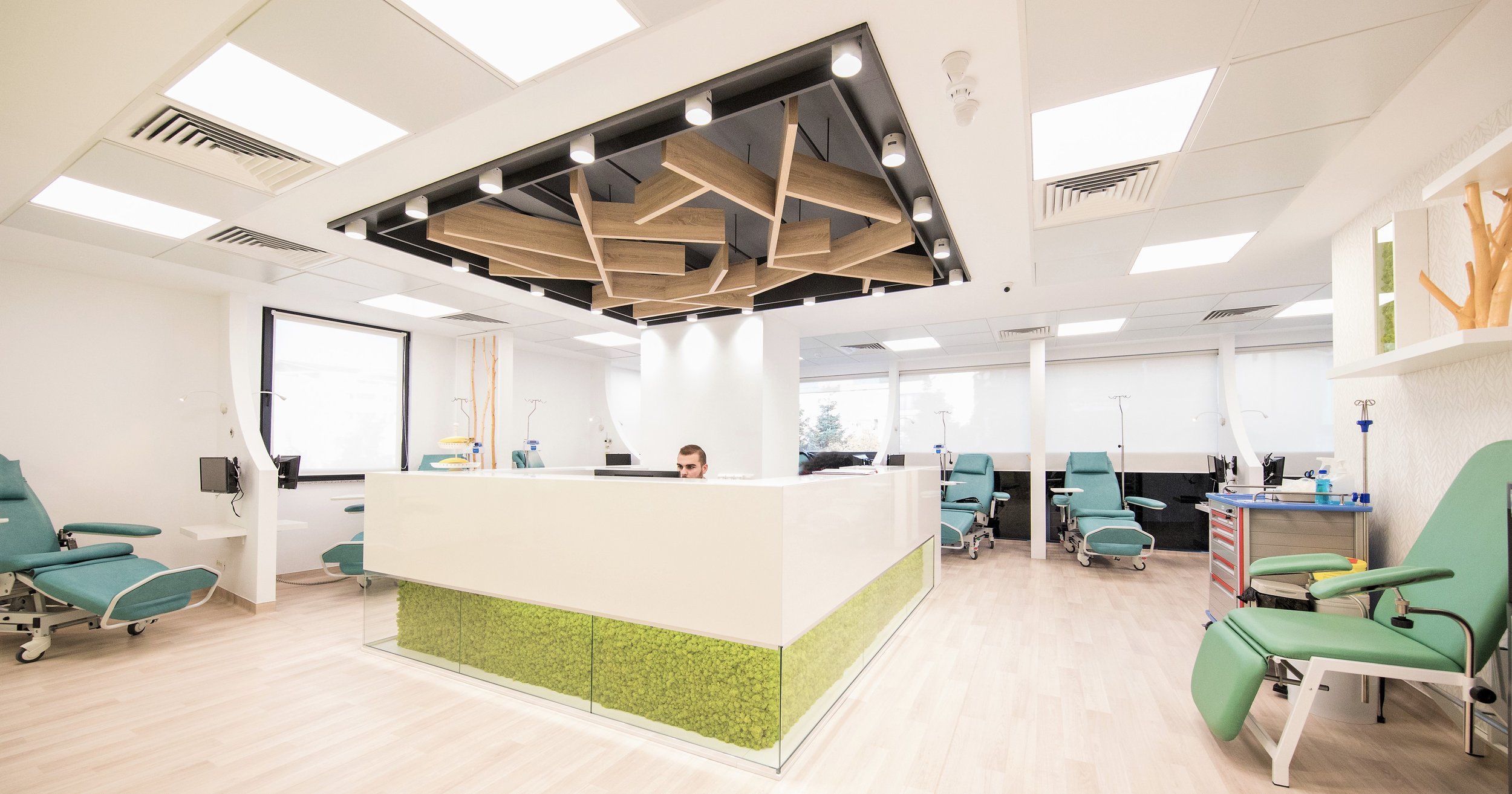 A modern hospital waiting area with teal medical recliners, IV stands, and a reception desk in the center. The ceiling has a decorative wooden panel with lighting, and large windows provide natural light.