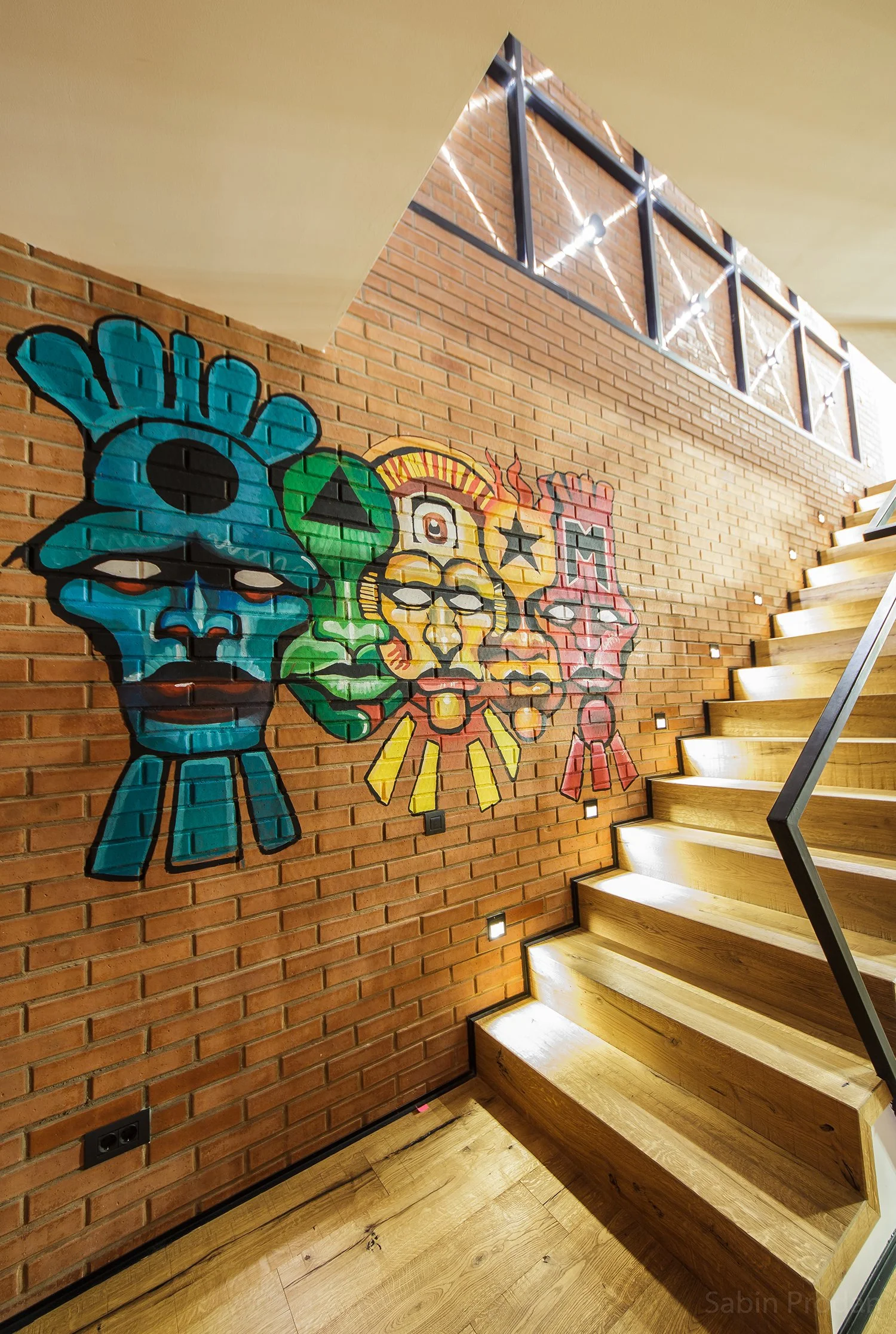Colorful mural of stylized faces and symbols painted on a brick wall under a staircase, in a modern interior space.