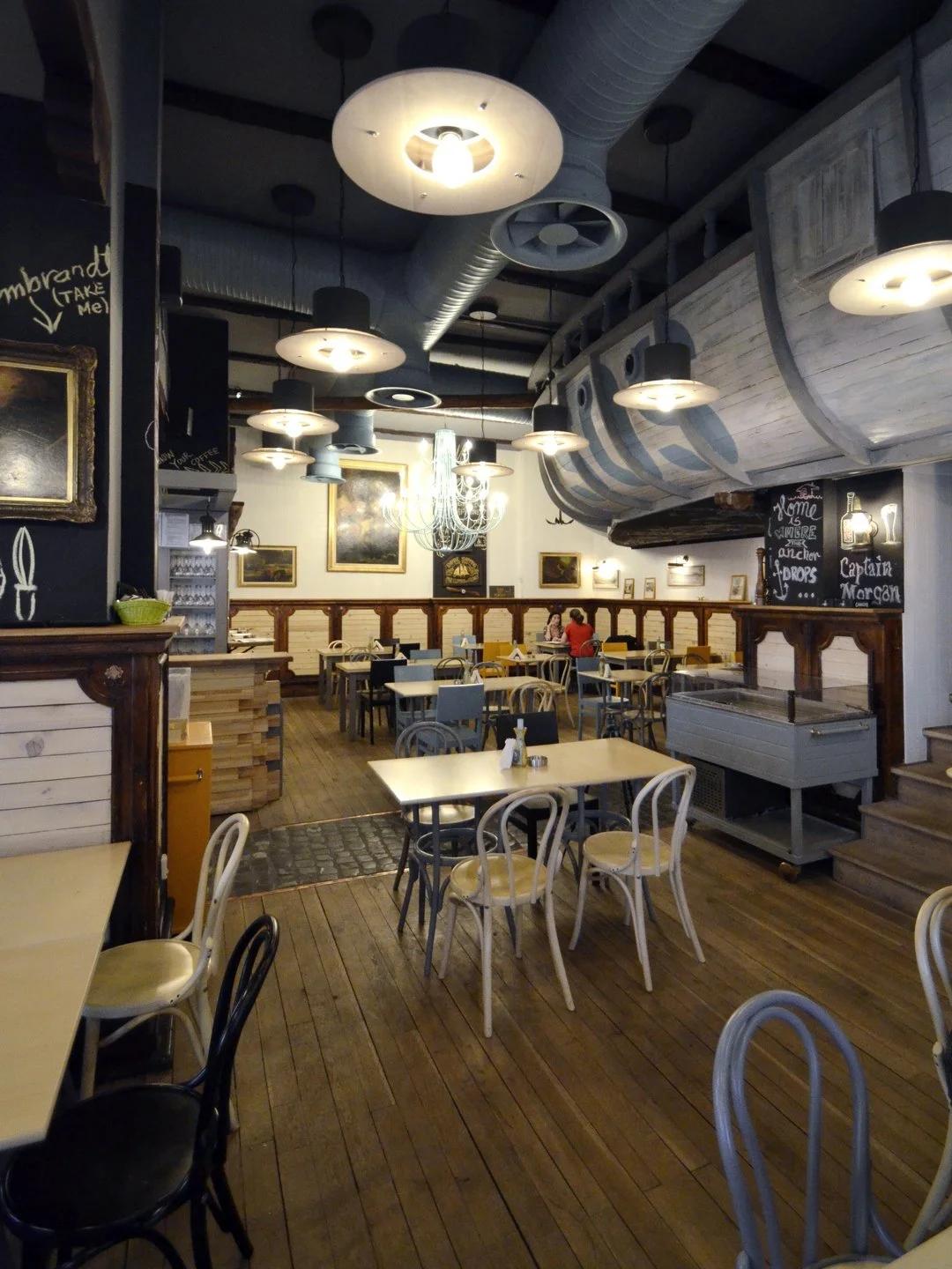 Interior of a cozy restaurant with wooden floors and black and beige chairs. Several tables are set with condiments. The ceiling has multiple round hanging lights and a chandelier, and a large ship's hull decoration is suspended overhead. Walls are decorated with framed paintings and there are a few patrons seated in the back.