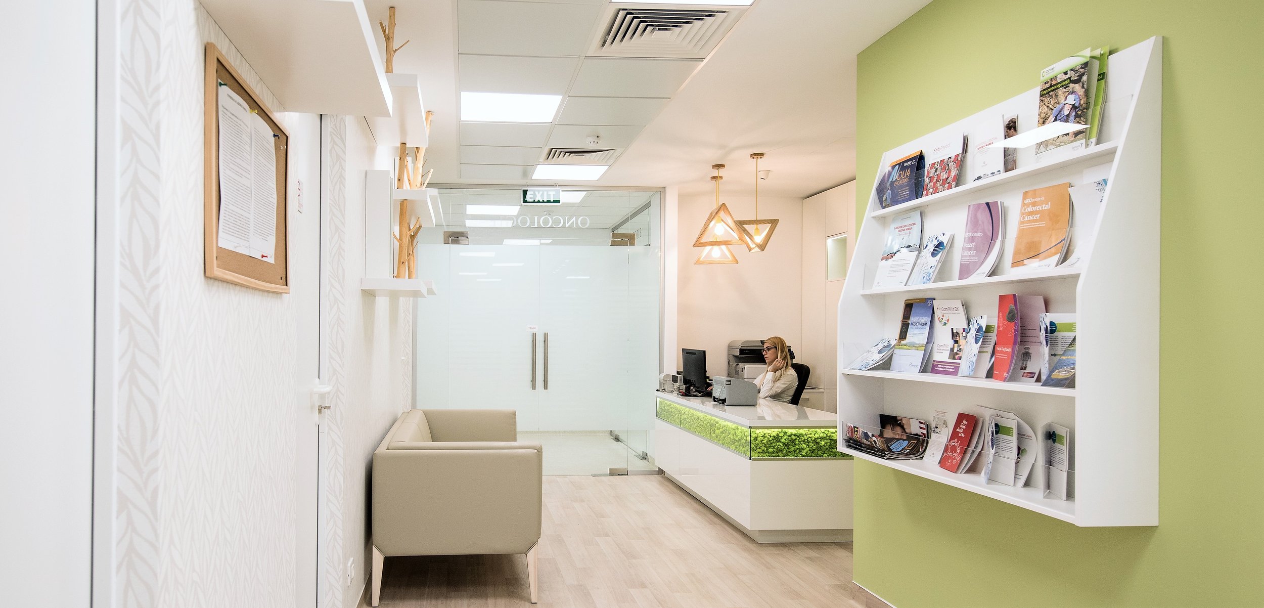 Reception area of a medical office with a woman sitting at a desk, a green accent wall with a white magazine rack, a beige couch, and decorative wall shelves.