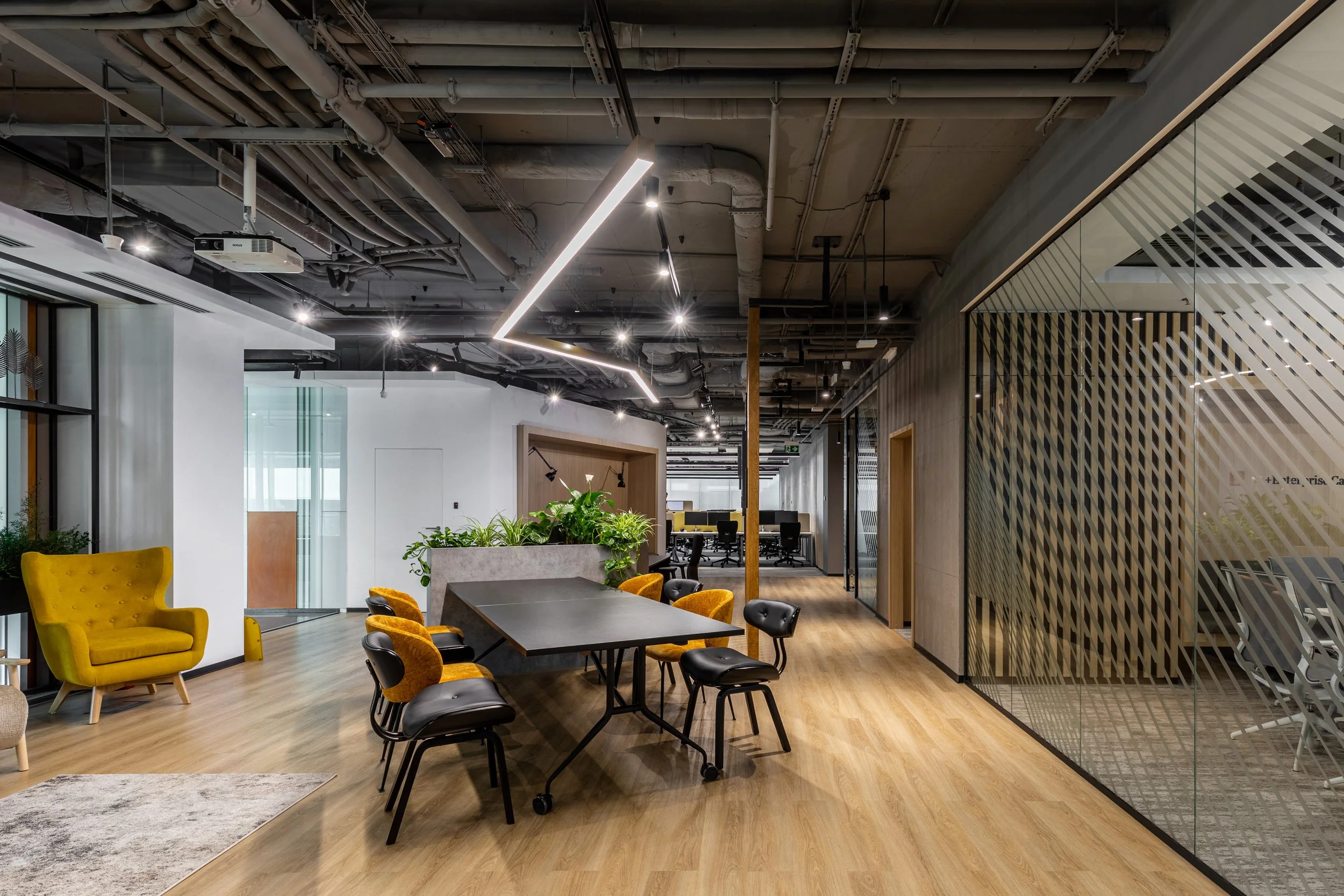 Modern open-concept office space with a wooden floor, black and yellow chairs around a black table, a yellow armchair, a large planter with green plants, and a meeting area with glass partitions.