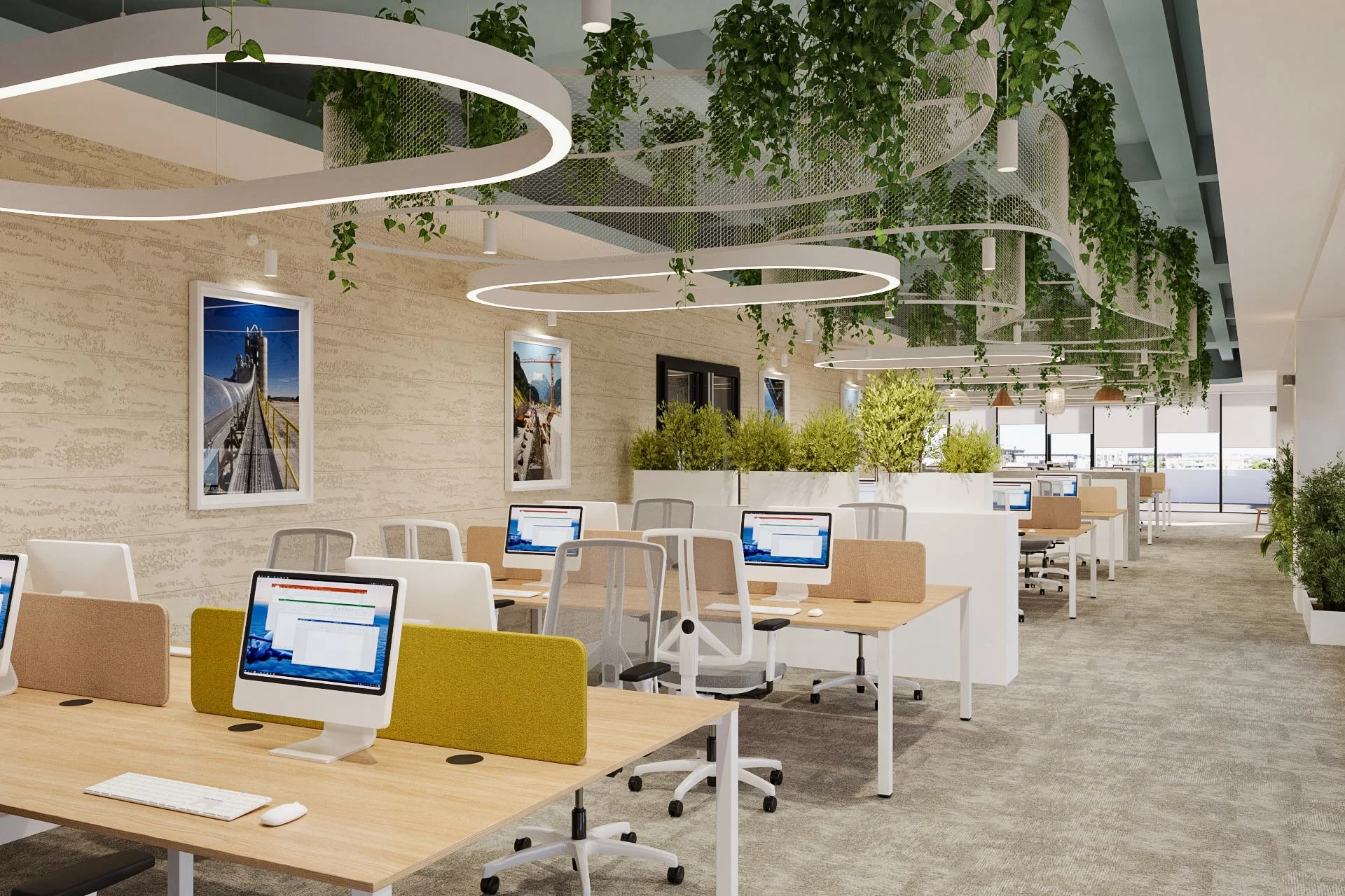 Modern open-plan office space with individual workstations equipped with desktop computers. The office features hanging circular light fixtures and abundant greenery including hanging plants and potted trees. Sunlight floods the room through large windows, and framed pictures adorn the beige stone walls.