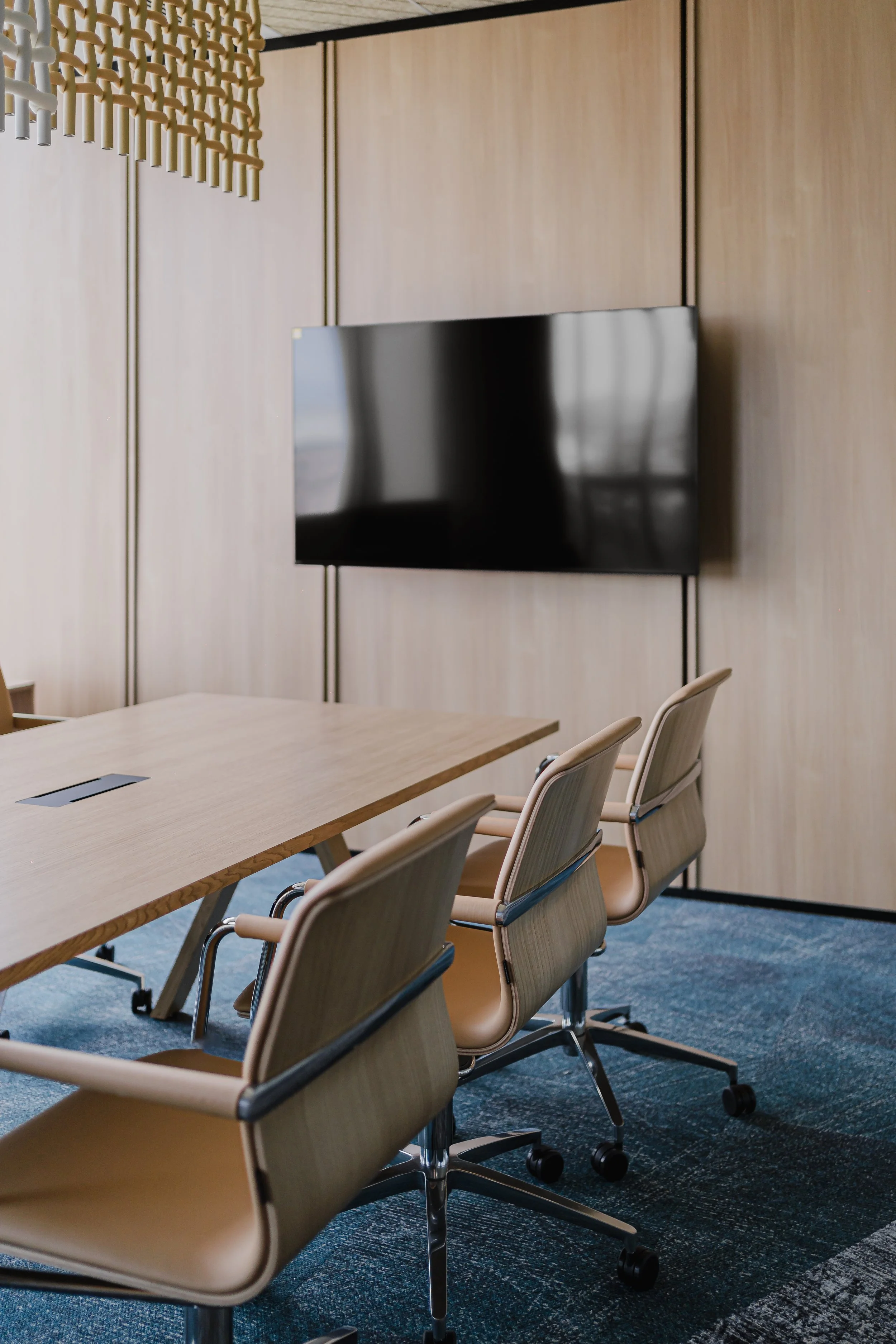 A modern conference room with a wooden table, beige chairs, a wall-mounted TV, and a wooden wall panel.