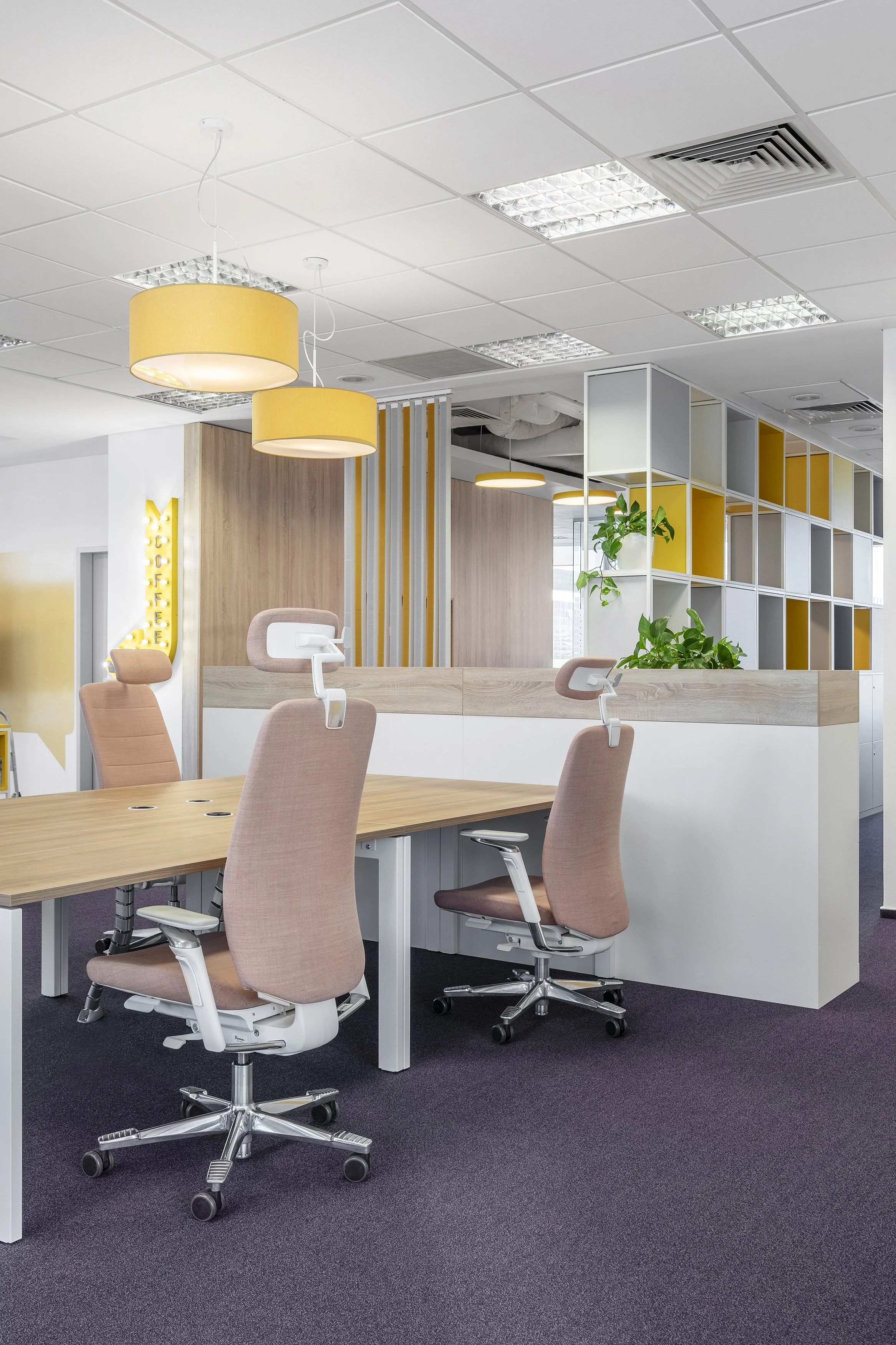 Modern office workspace with pink office chairs, wooden desk, hanging yellow lamps, and a decorative partition with plants.