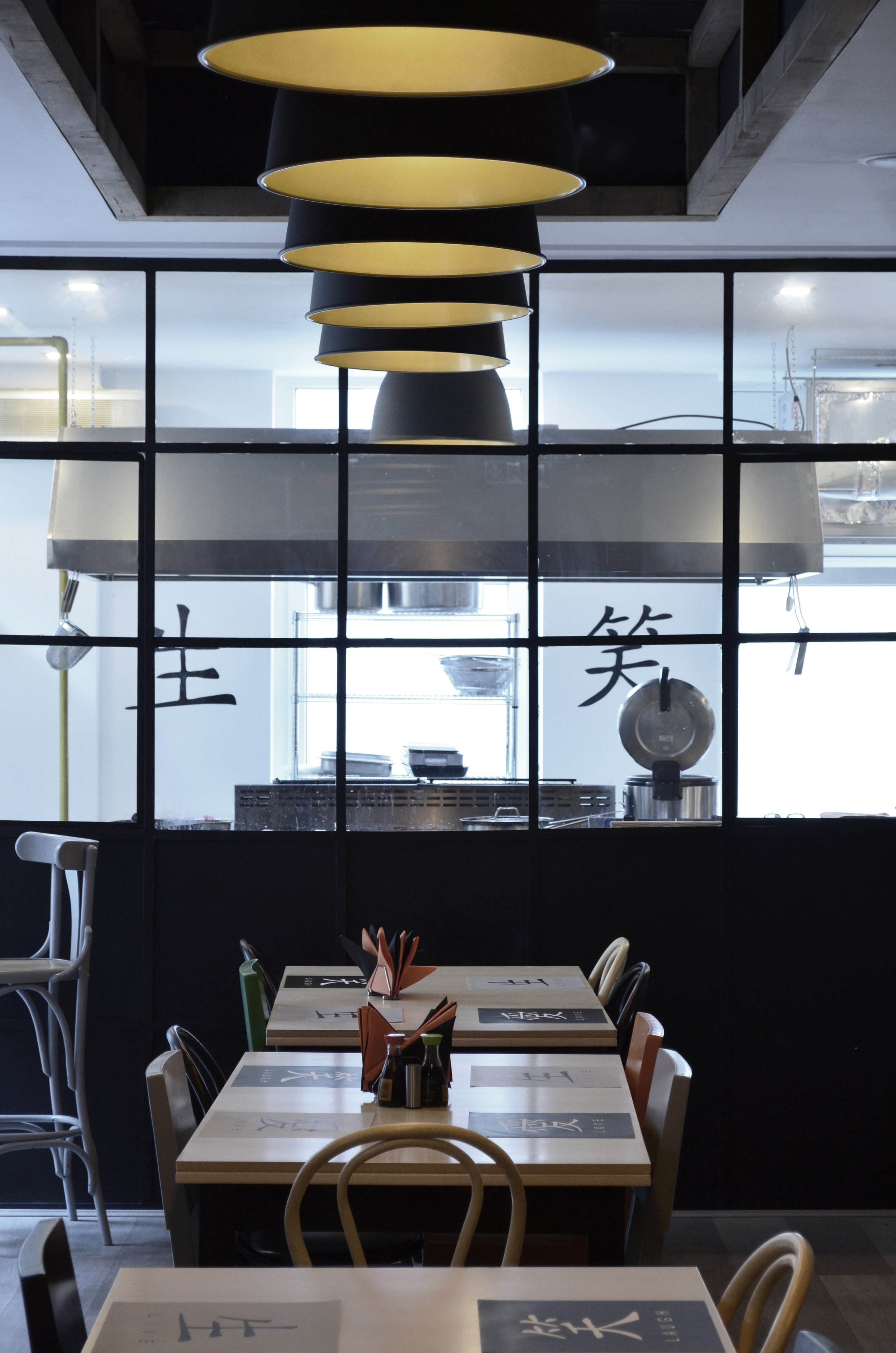 View of a restaurant's interior, including a table set with menus, a wall with Chinese characters, and a view into the kitchen through a glass partition with modern lighting overhead.