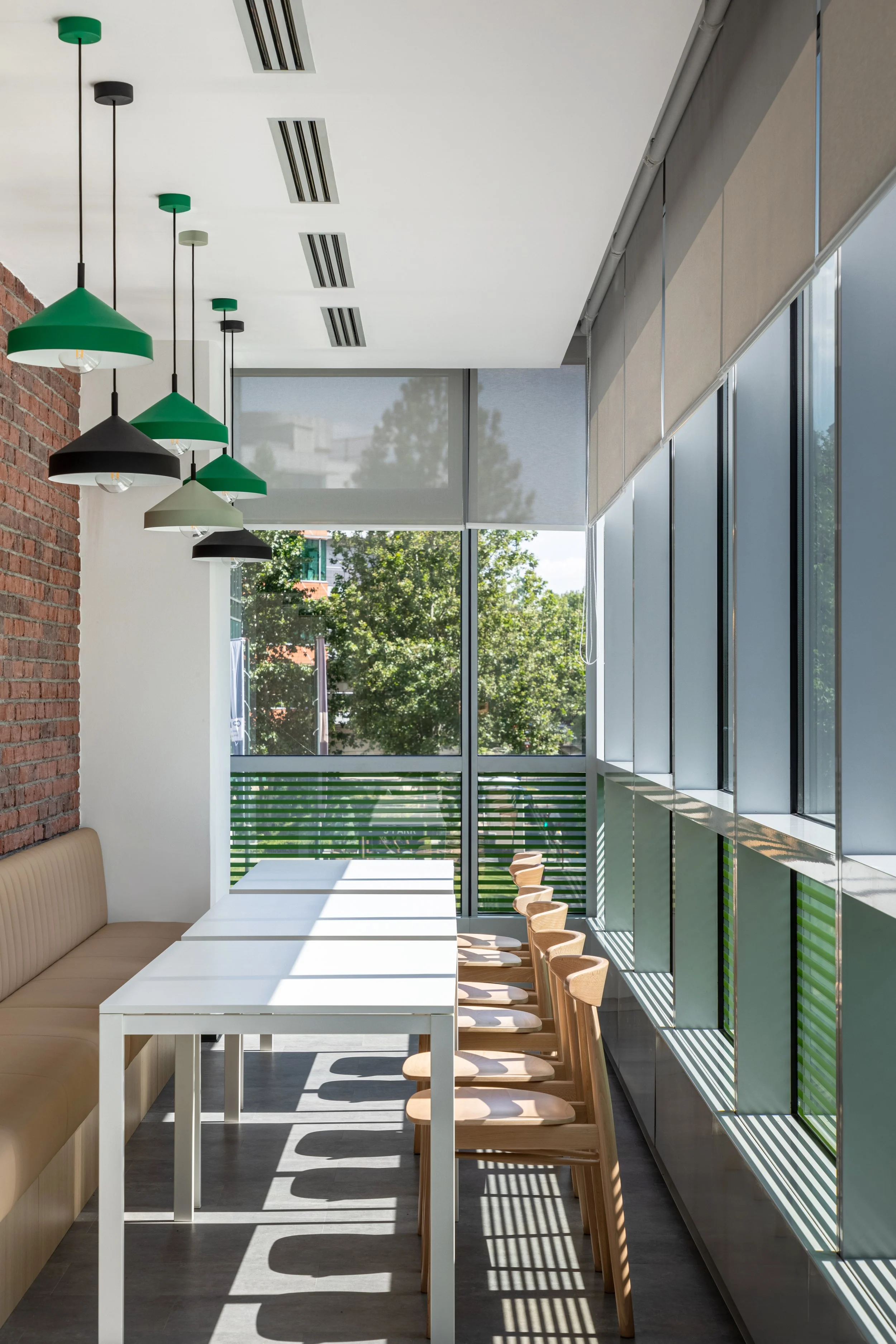 Modern dining area with white table, wooden chairs, beige bench, hanging green and black pendant lights, large windows with roller shades, outdoor trees visible.