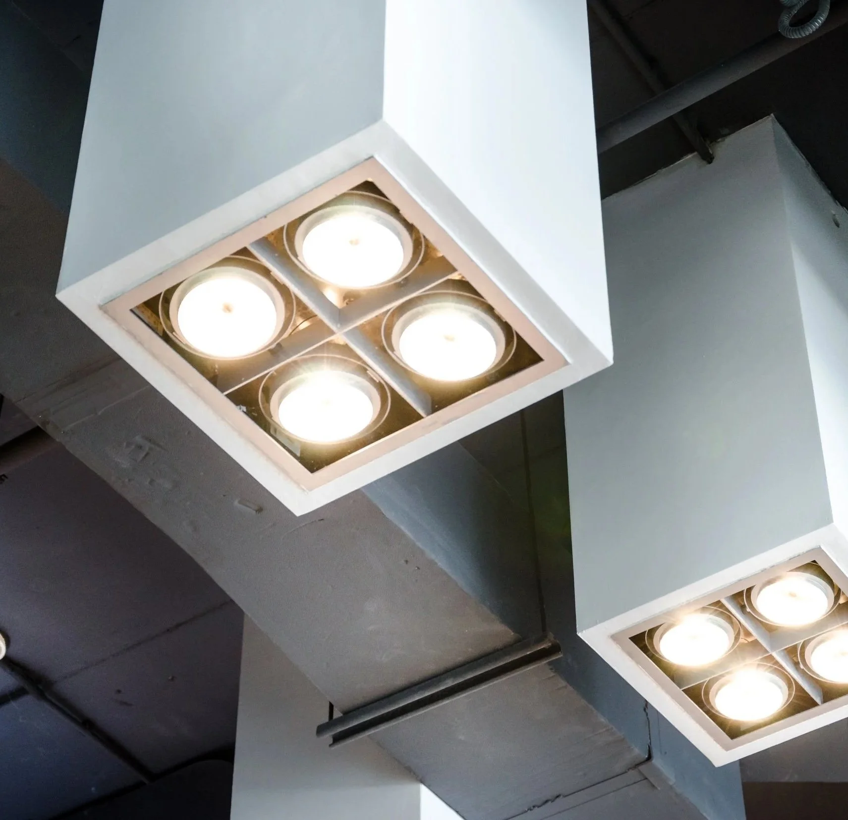 Modern ceiling lights with four round bulbs each in square white fixtures hanging from a ceiling with exposed ductwork.
