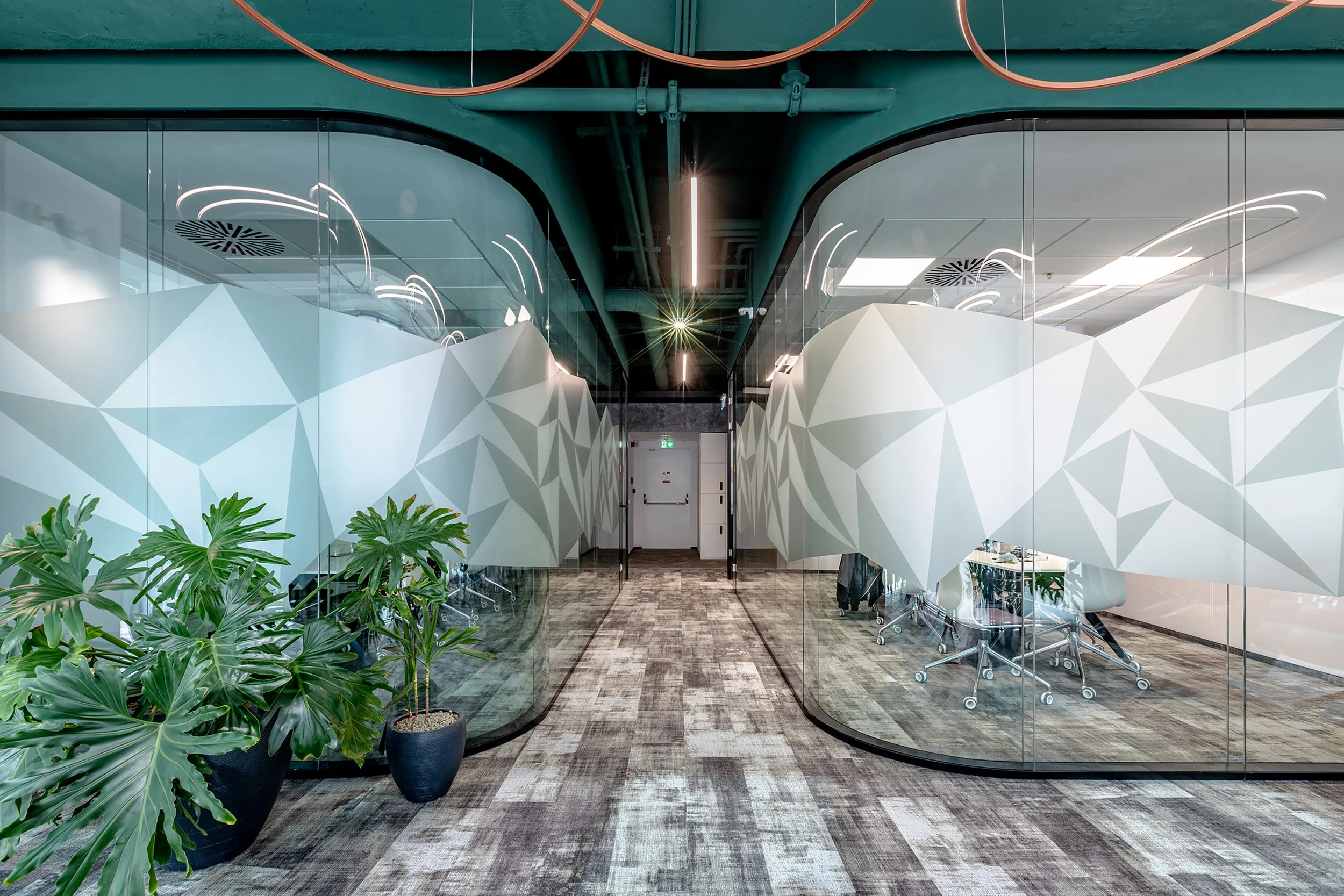 Modern office conference room with glass walls decorated with geometric white patterns, potted green plants, and rolling office chairs.