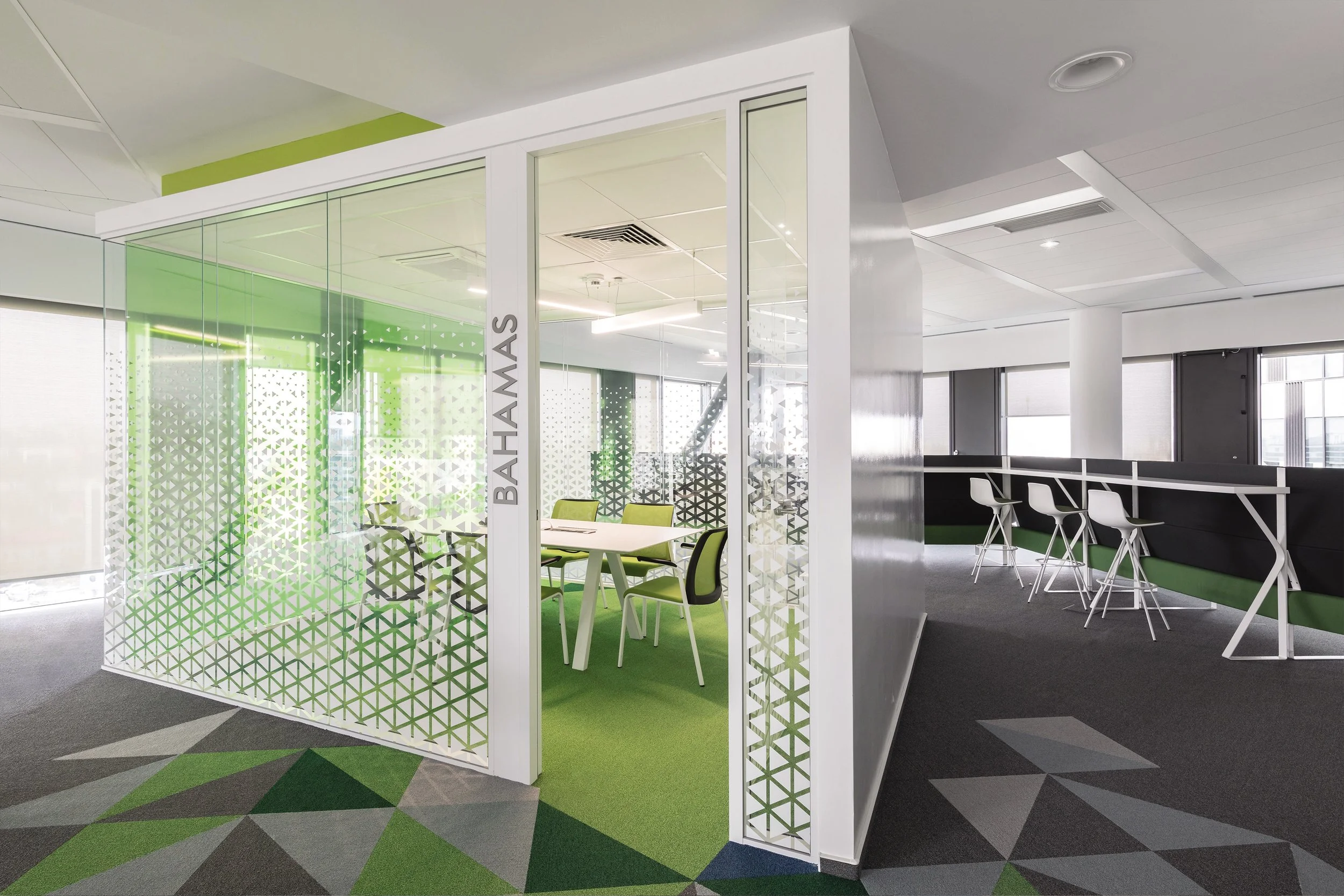 Modern office lounge area with glass enclosed meeting room labeled 'BAHAWAS', green and gray geometric patterned carpet, and white barstools along a high counter.