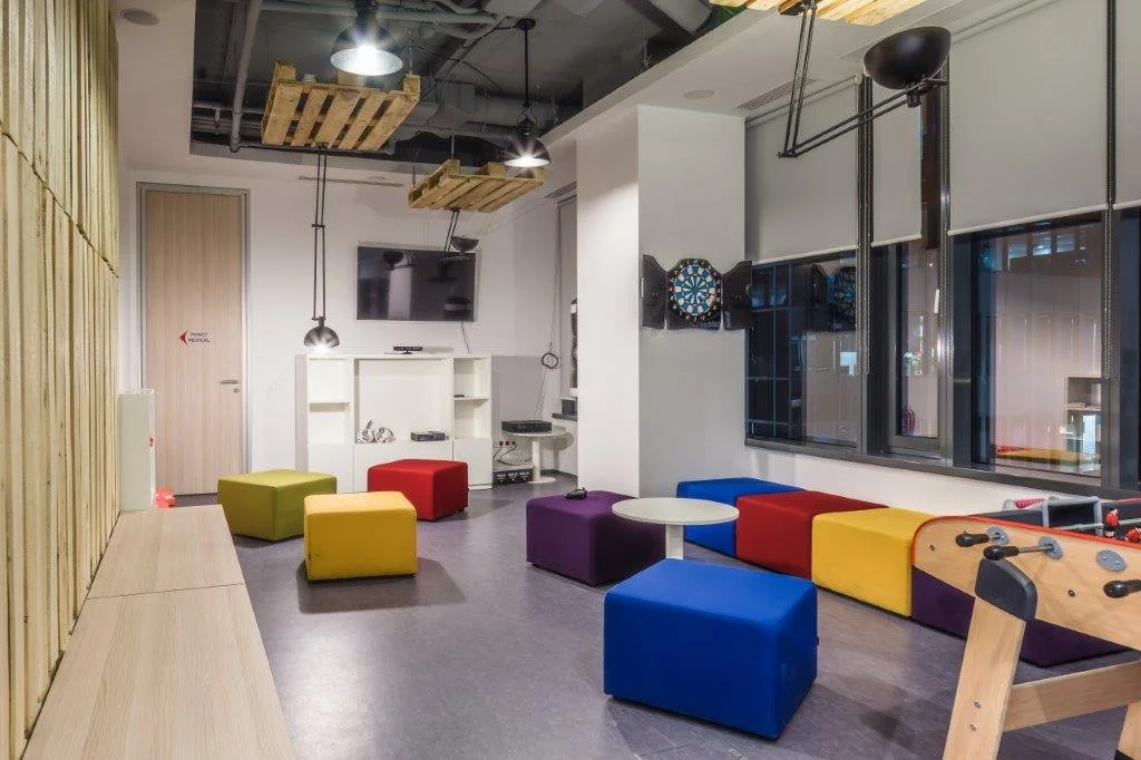 A colorful indoor recreational room with multicolored ottomans, a small round table, darts on the wall, a pool table, and a large window.
