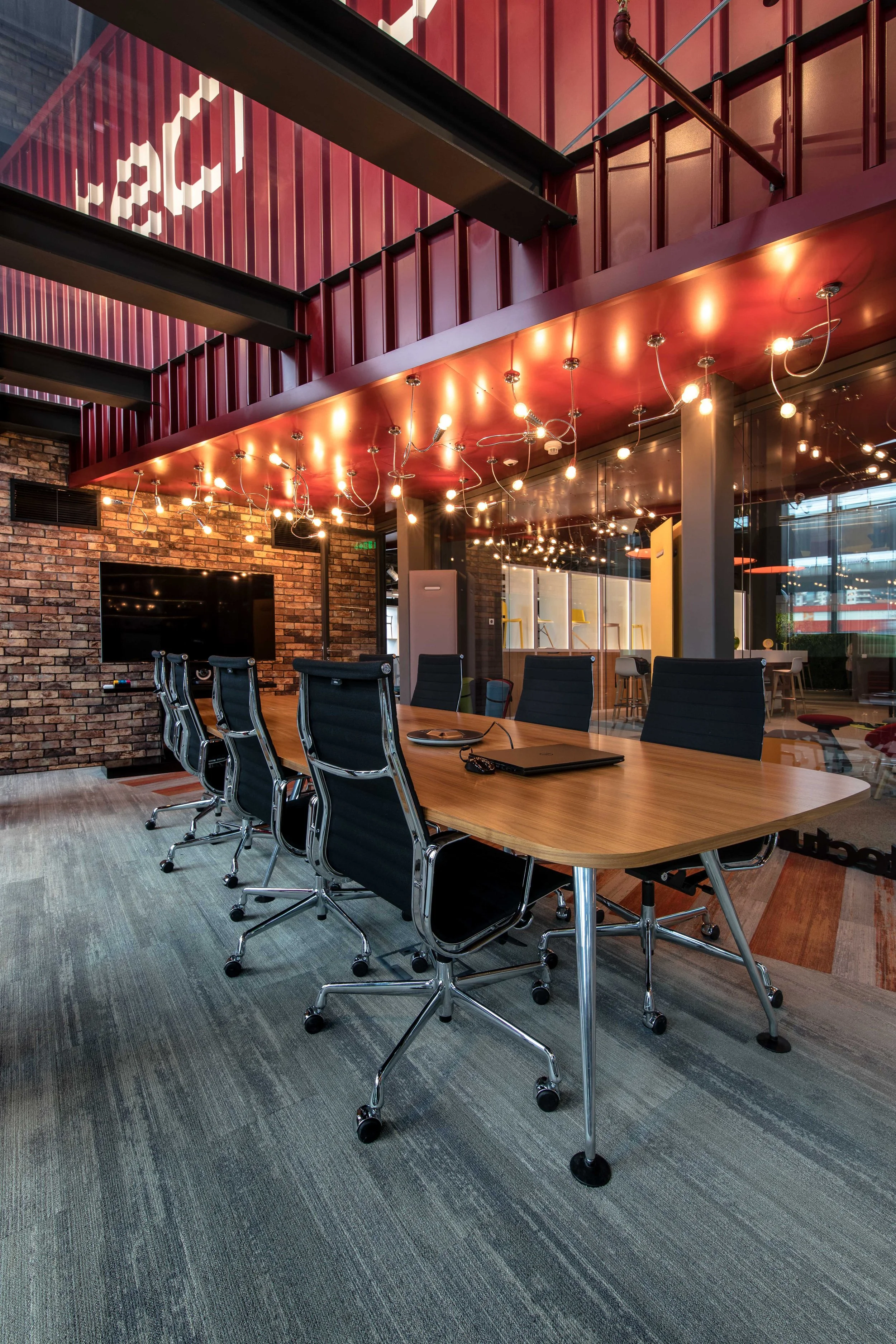 Modern conference room with a large wooden table, black leather chairs, brick wall, flat-screen TV, and string lights hanging from a red ceiling.