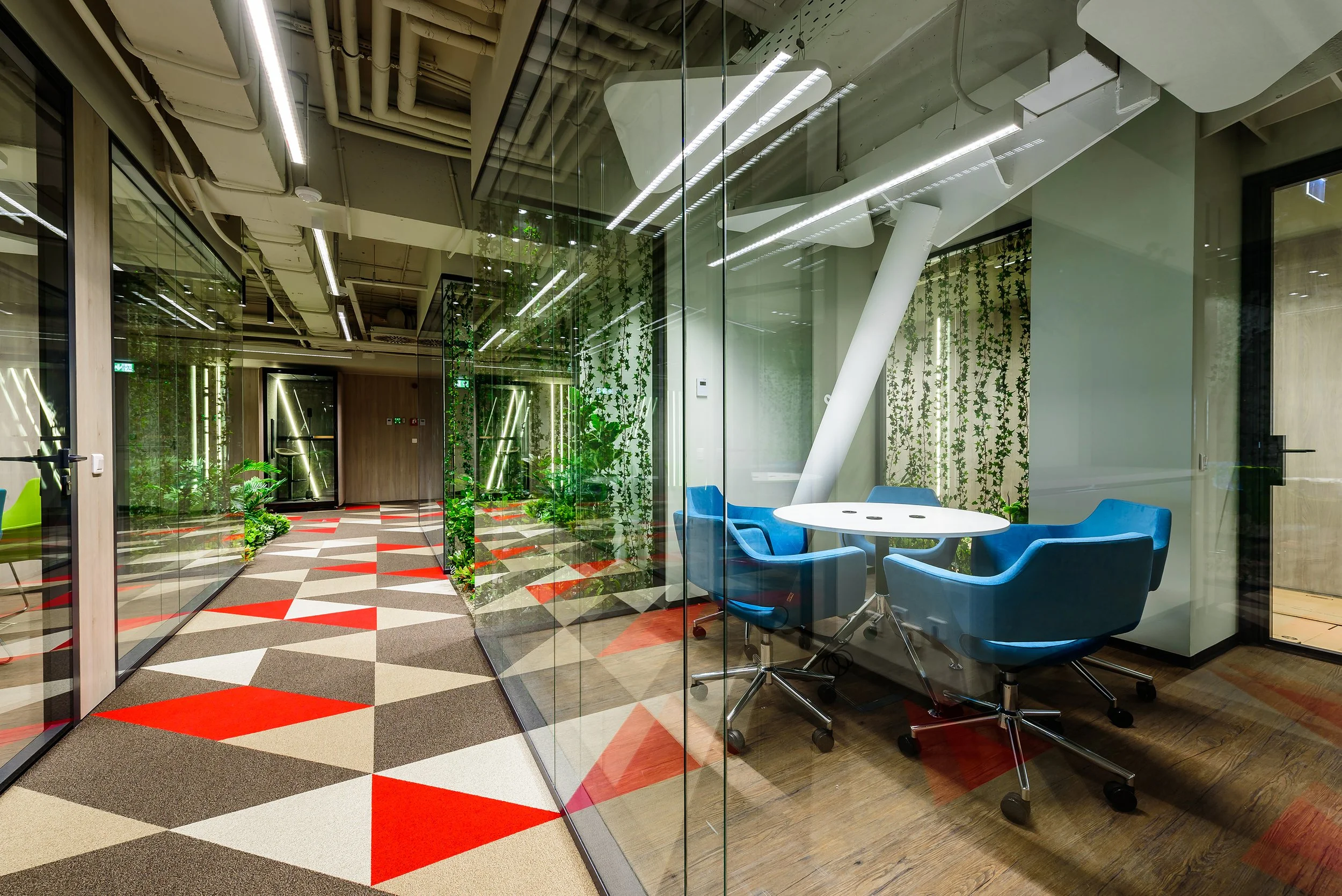 Modern office interior with glass walls, a small round table with four blue chairs, green plants, and decorative lighting fixtures.