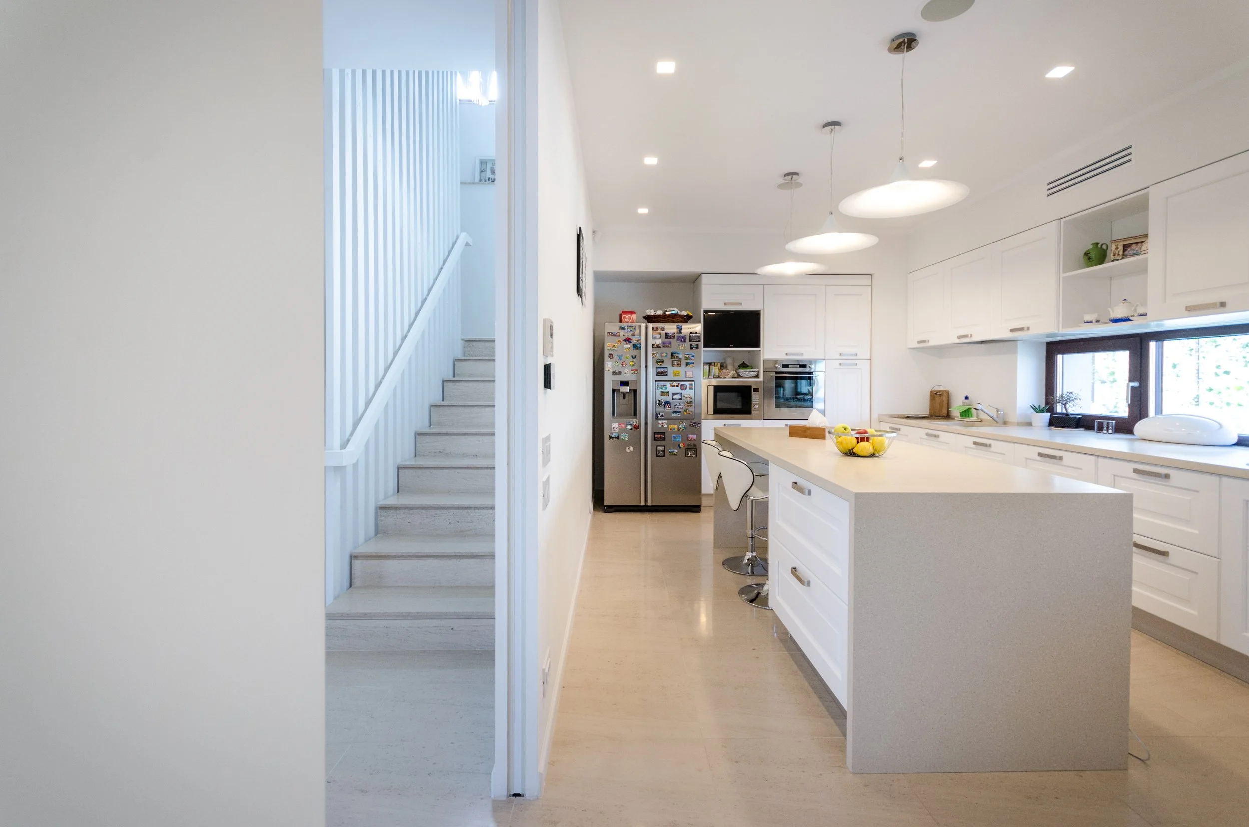 Modern kitchen with white cabinets, a large island with a fruit bowl, stainless steel refrigerator, oven, microwave, and a window above the sink. String lights hang from the ceiling, and stairs are visible on the left.