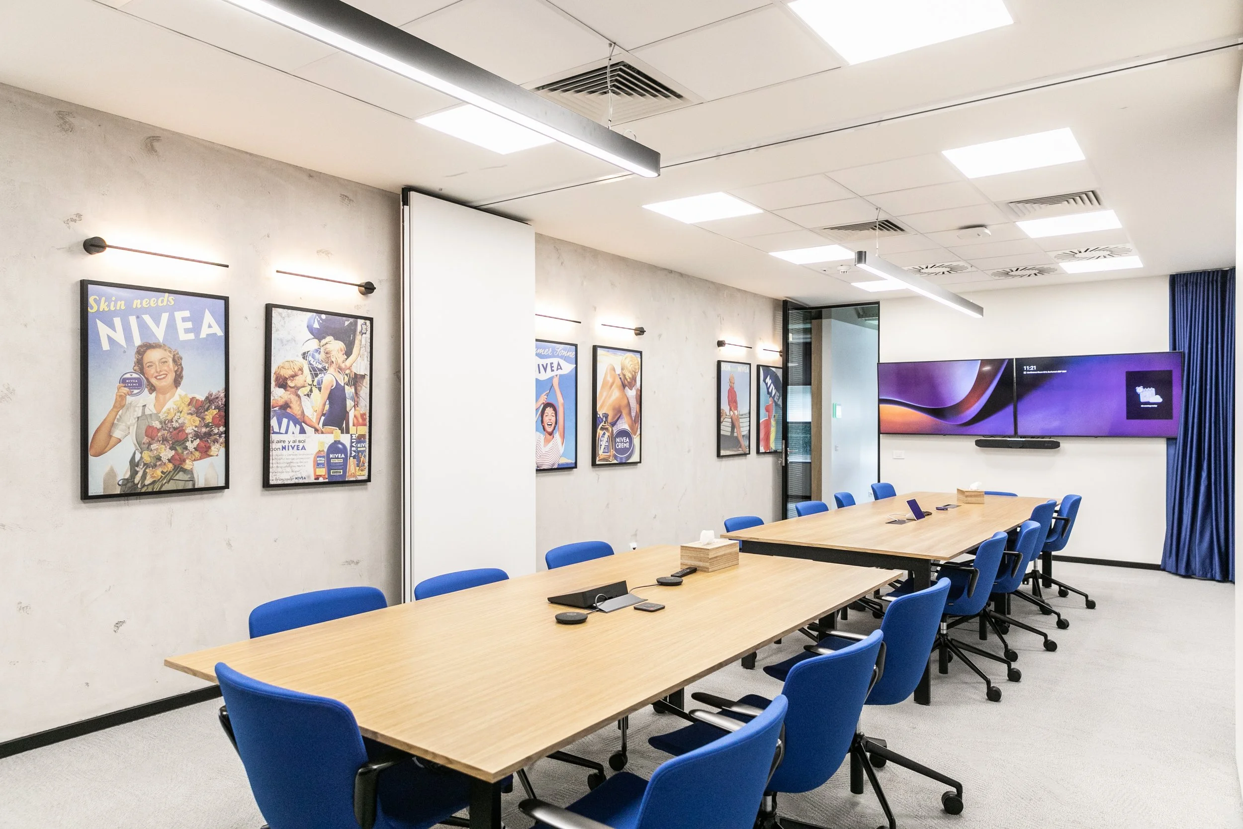 A modern conference room with a large wooden table surrounded by blue chairs. There are framed vintage advertisements on the wall promoting Nivea skincare, and a large digital screen on the far wall displaying a purple and black abstract background. The room has a light-colored carpet, white walls, and ceiling with bright lighting.